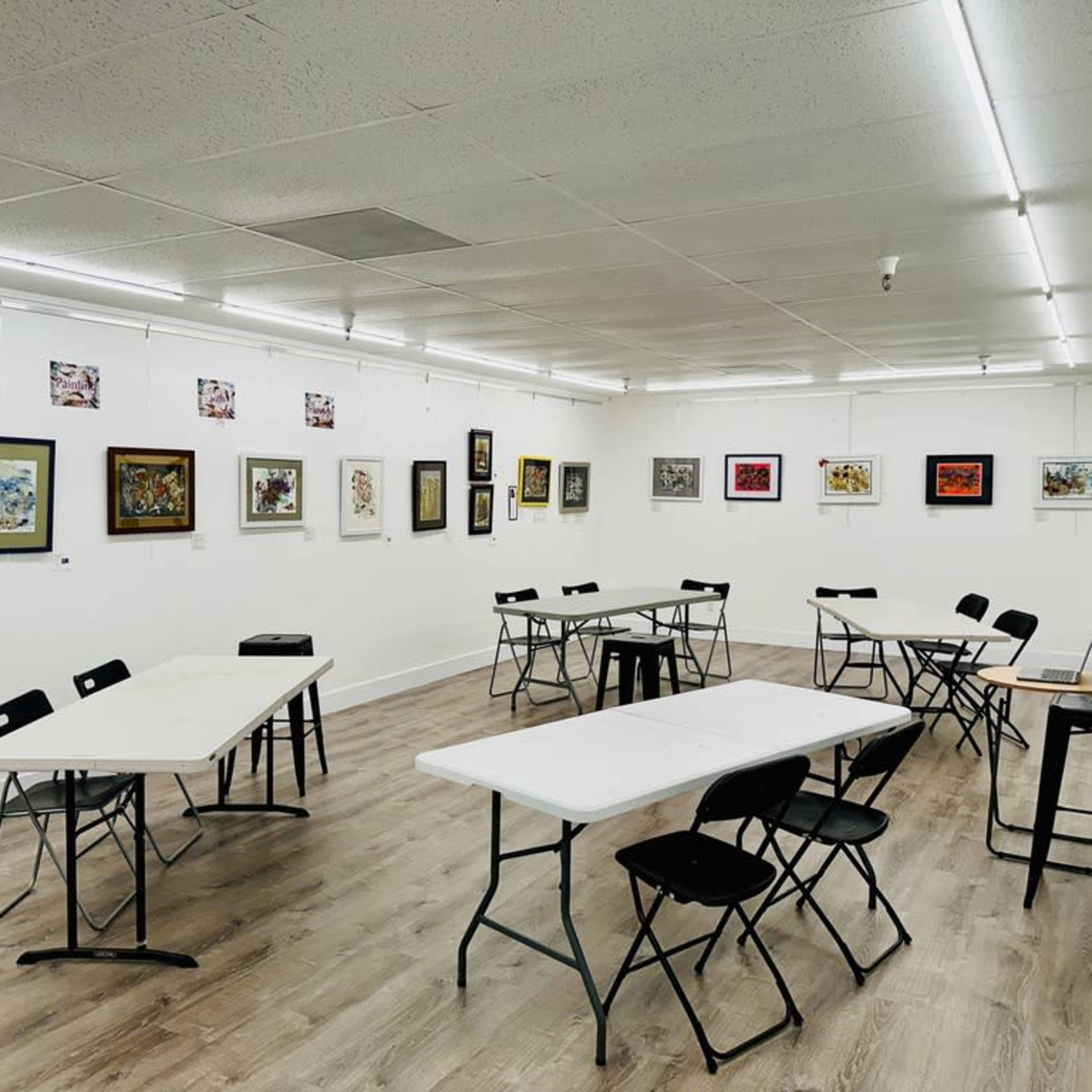An art gallery with several framed artworks displayed on the walls and multiple tables and chairs arranged in the center.