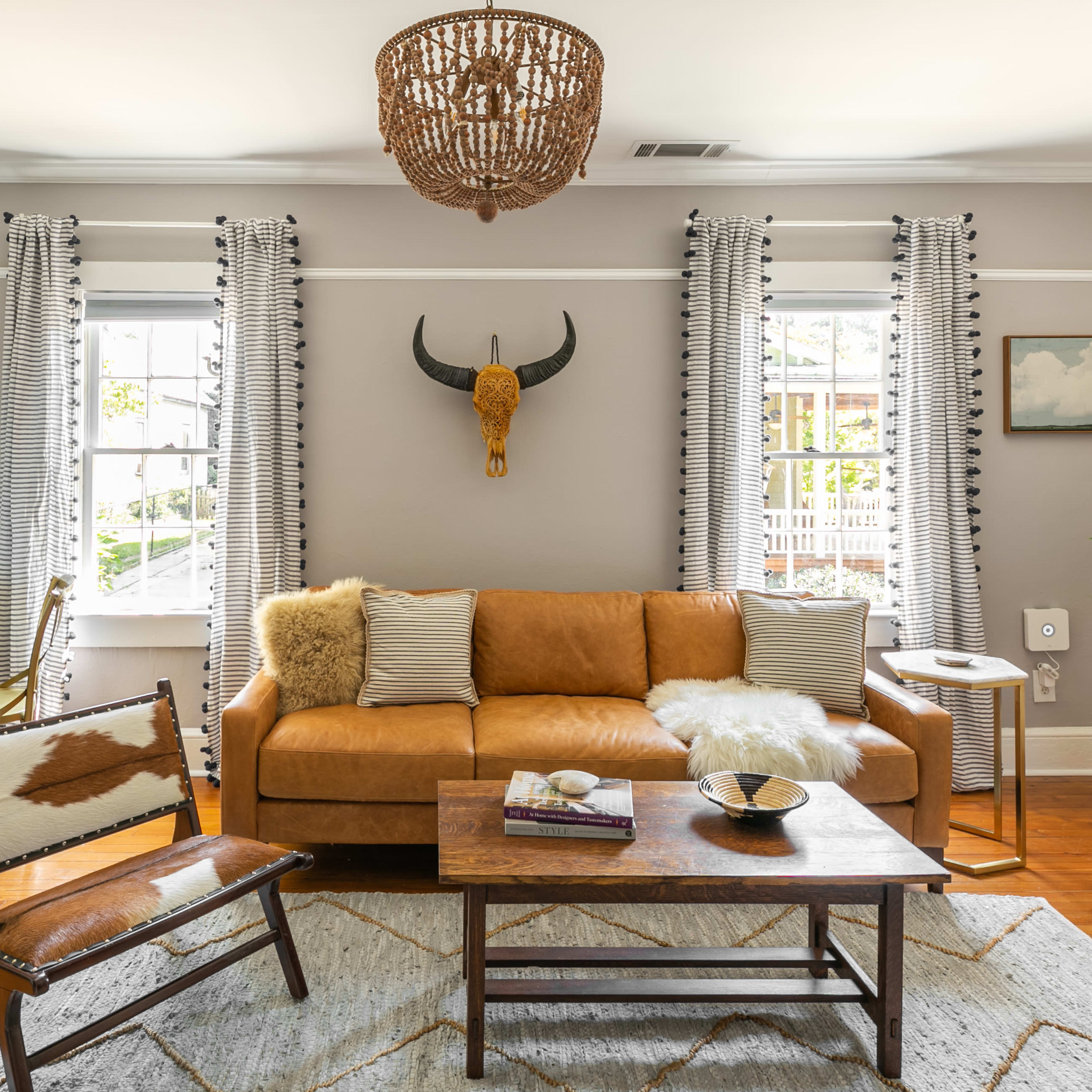 The living room features a brown leather sofa, a wooden coffee table, and a vintage chair, with large windows dressed in striped curtains and a decorative bull skull mounted on the wall.