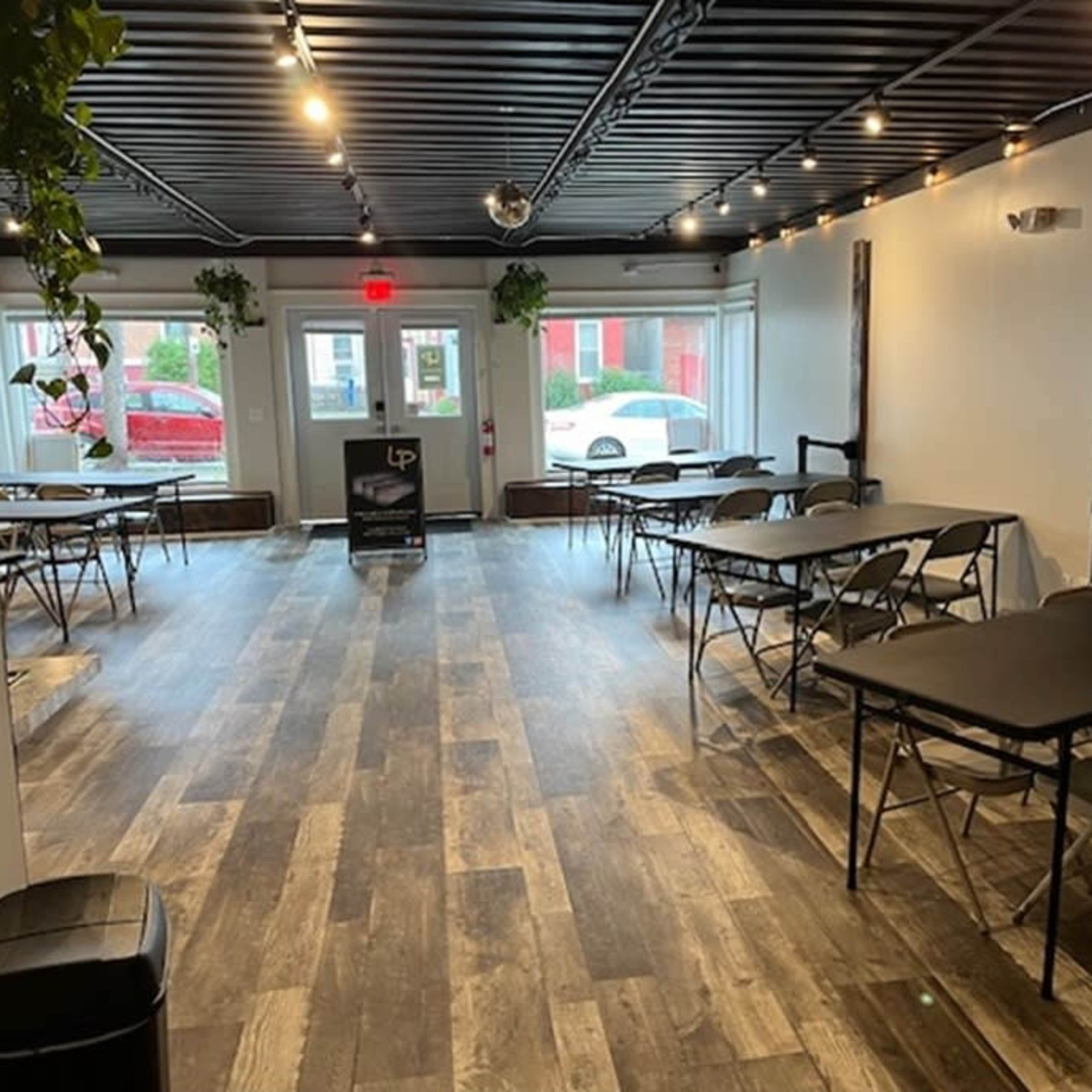 A spacious room with several rows of folding tables and chairs, illuminated by ceiling lights and featuring wooden flooring.