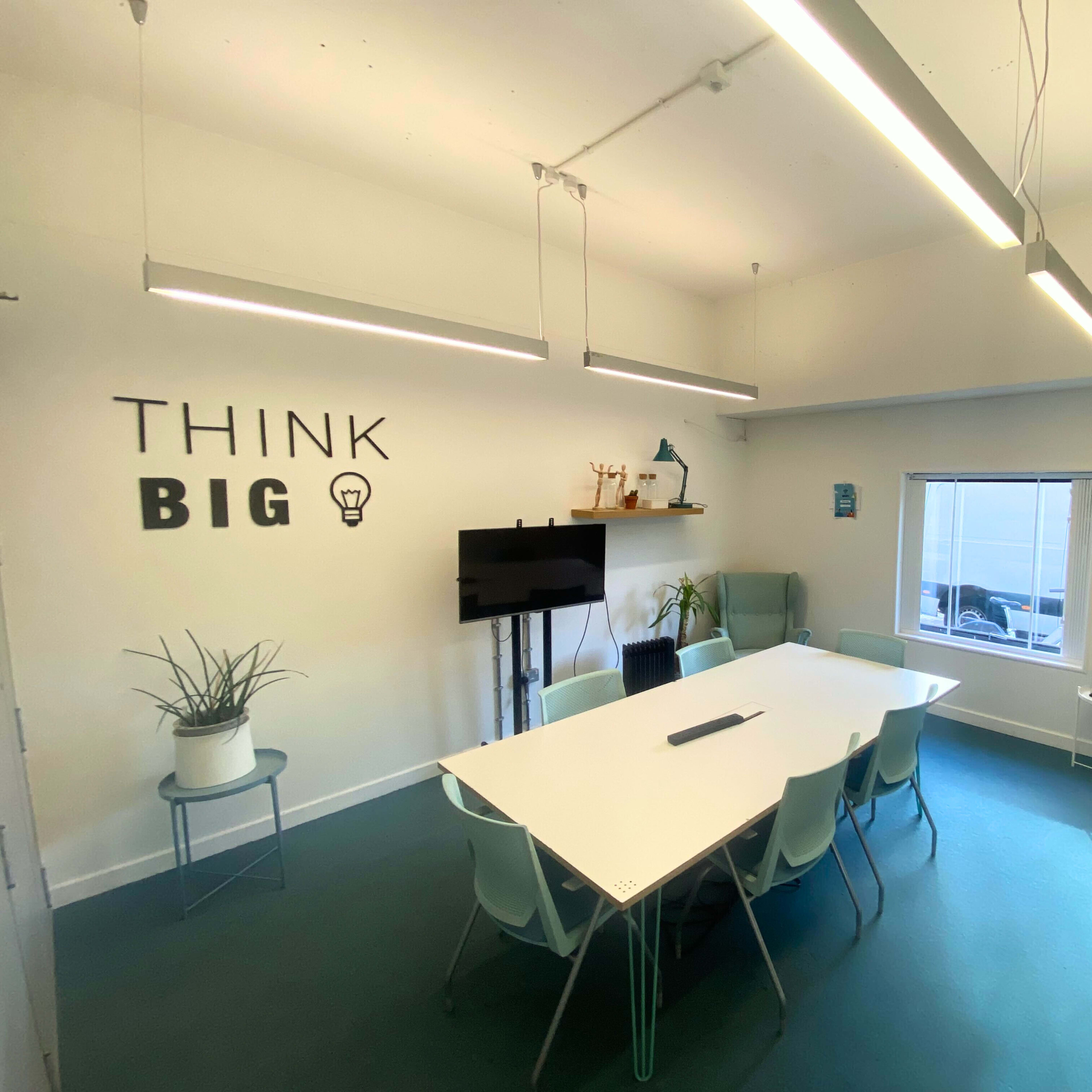 A well-lit meeting room with a large white table surrounded by light-colored chairs, a television on the wall, and the words "THINK BIG" displayed decoratively.