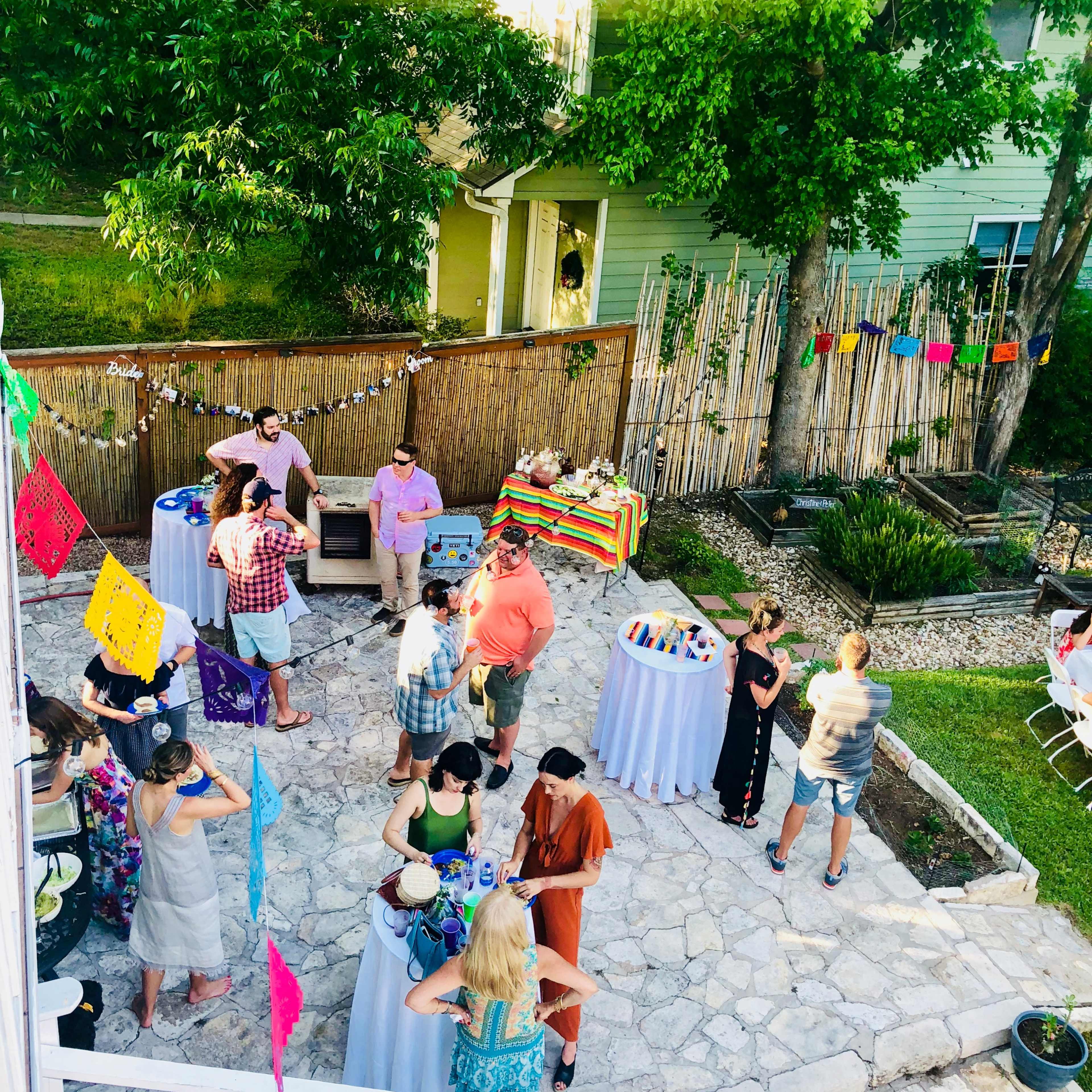 A diverse group of people gathers around tables adorned with colorful items at an outdoor party in a garden setting.