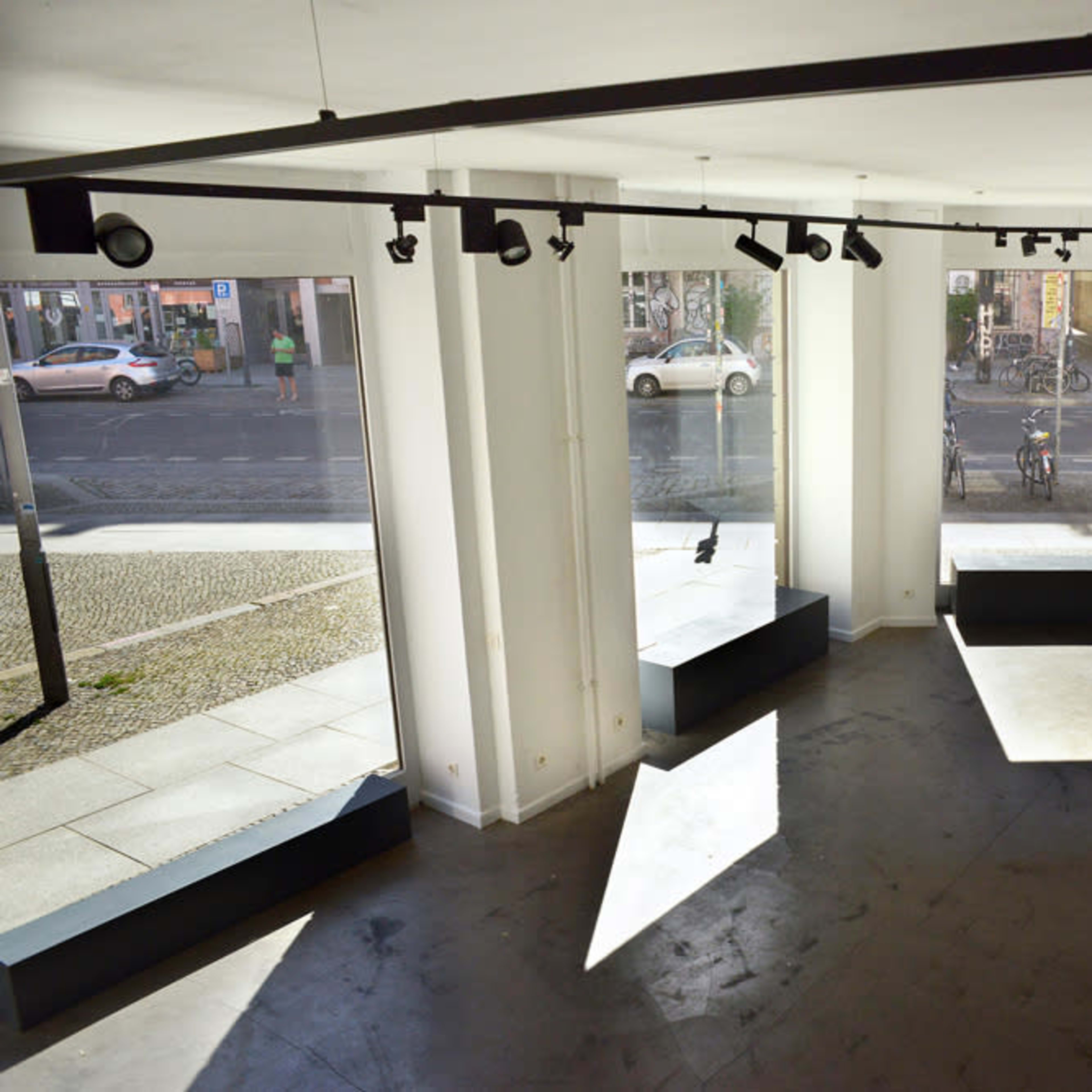 An empty art gallery with large windows, track lighting, and white walls, overlooking a street.