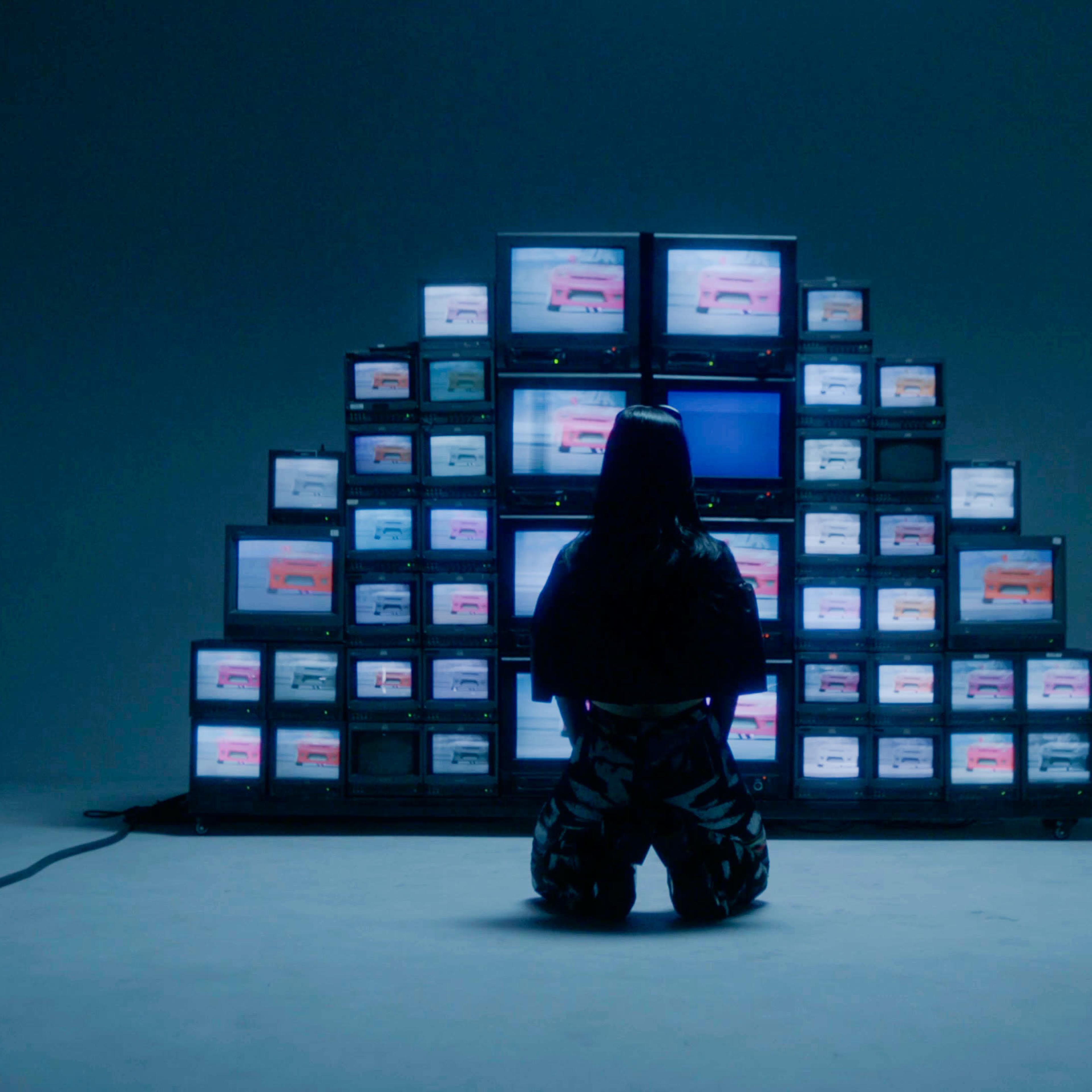 A person kneels in front of a large arrangement of multiple television screens displaying a series of images.