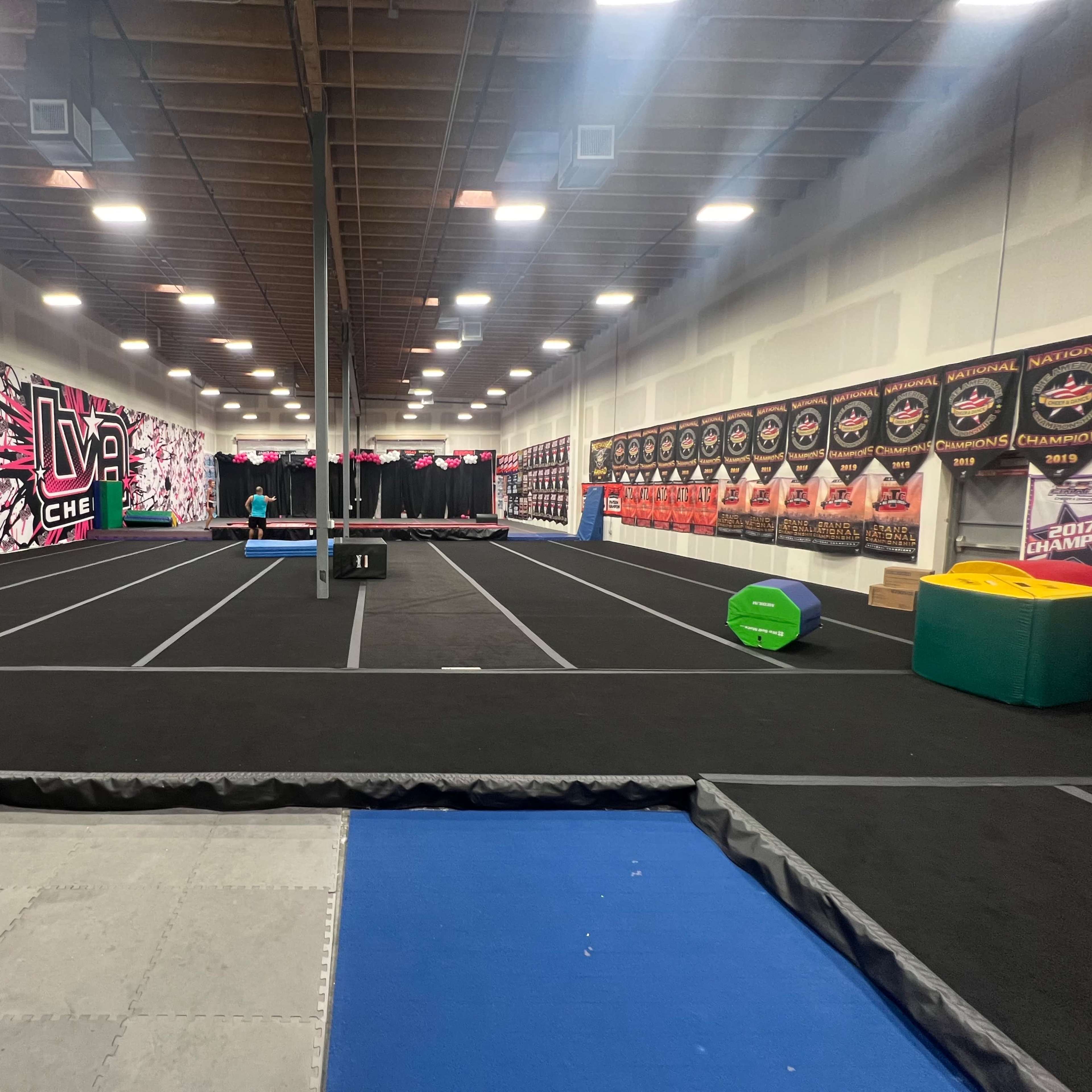 An indoor gymnastics facility with black flooring, framed by a colorful wall mural and lined with various training equipment along the sides.