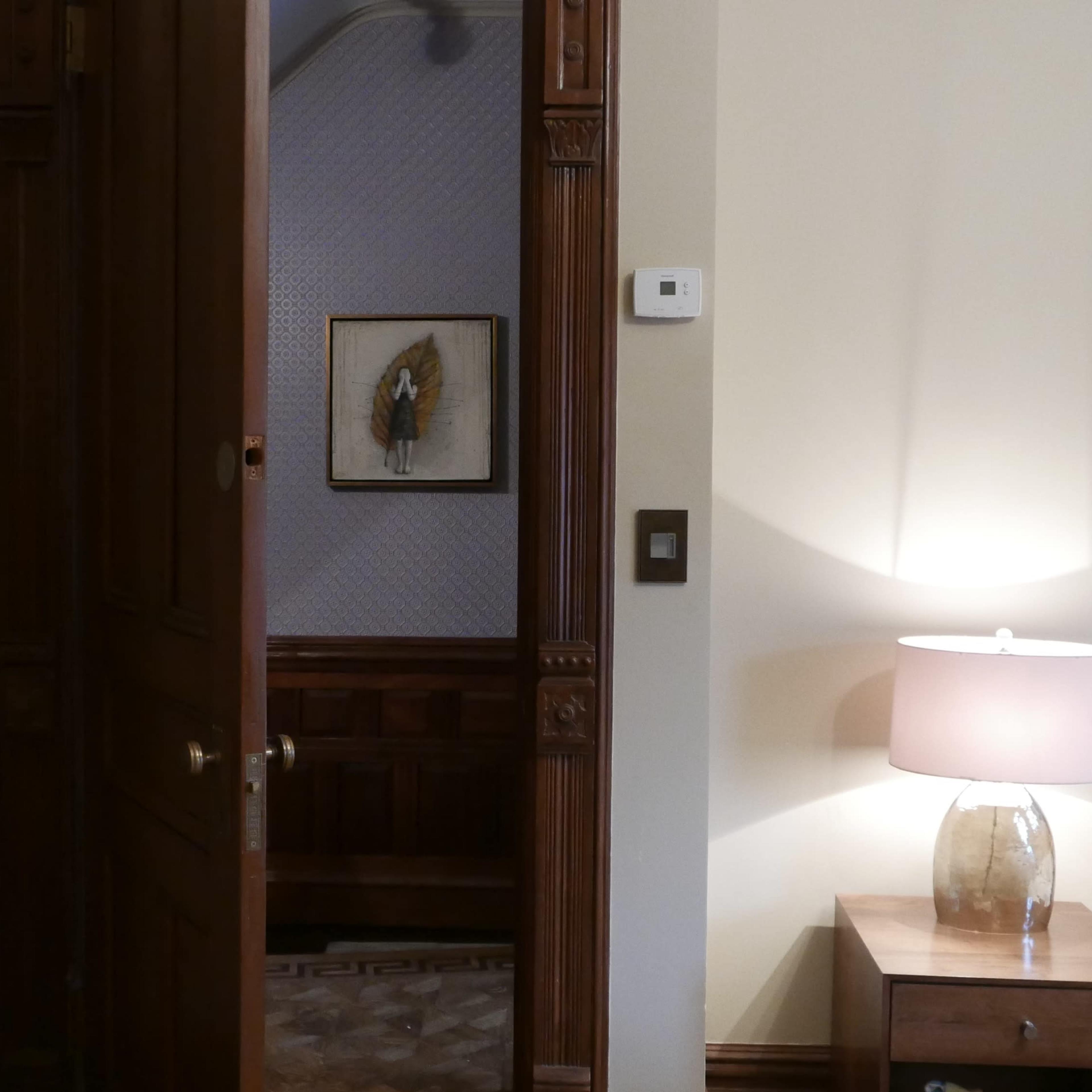 A wooden door partially open reveals a room with a bedside lamp and an artwork on the wall.