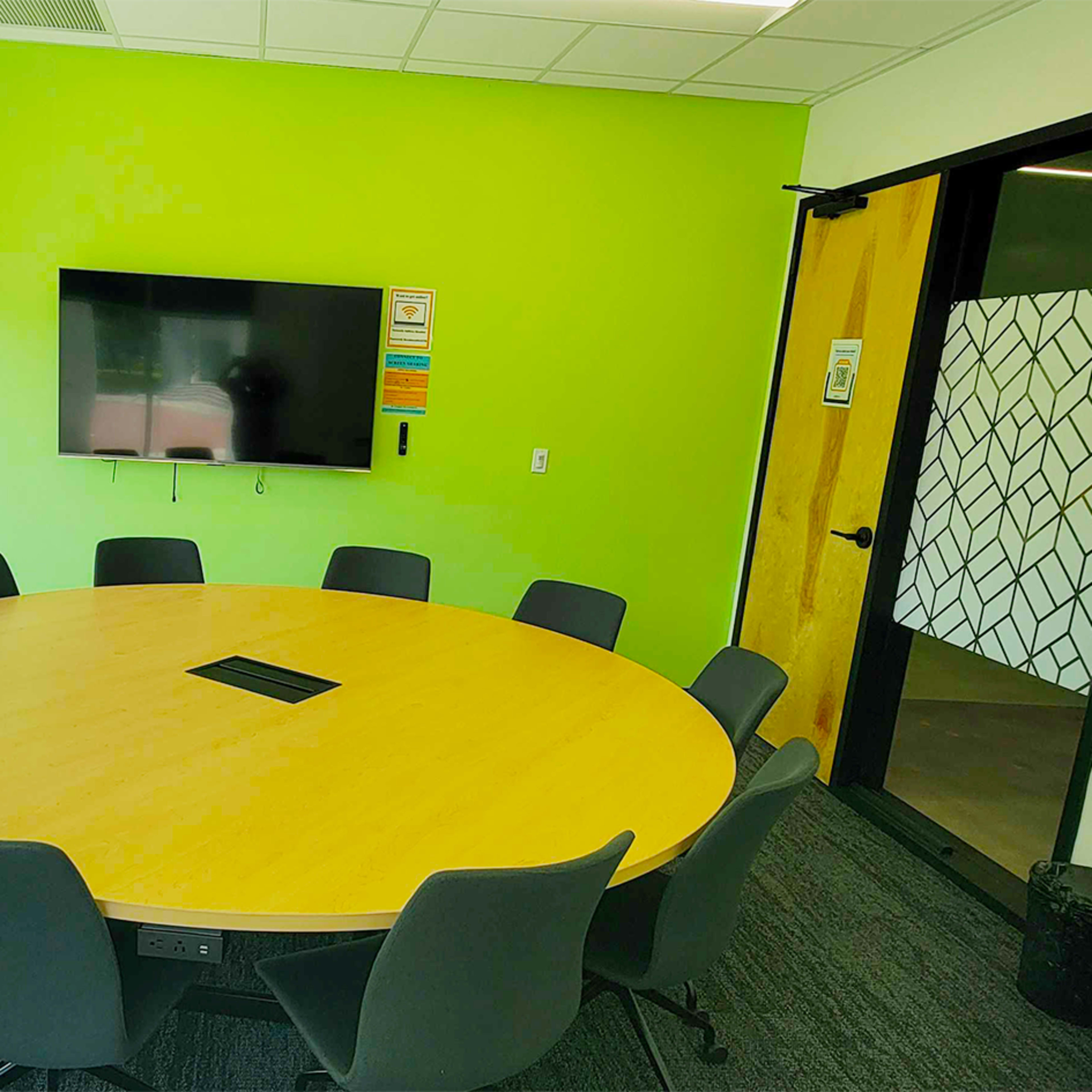 A round conference table surrounded by chairs is set in a brightly lit meeting room with a large window and a wall-mounted television.
