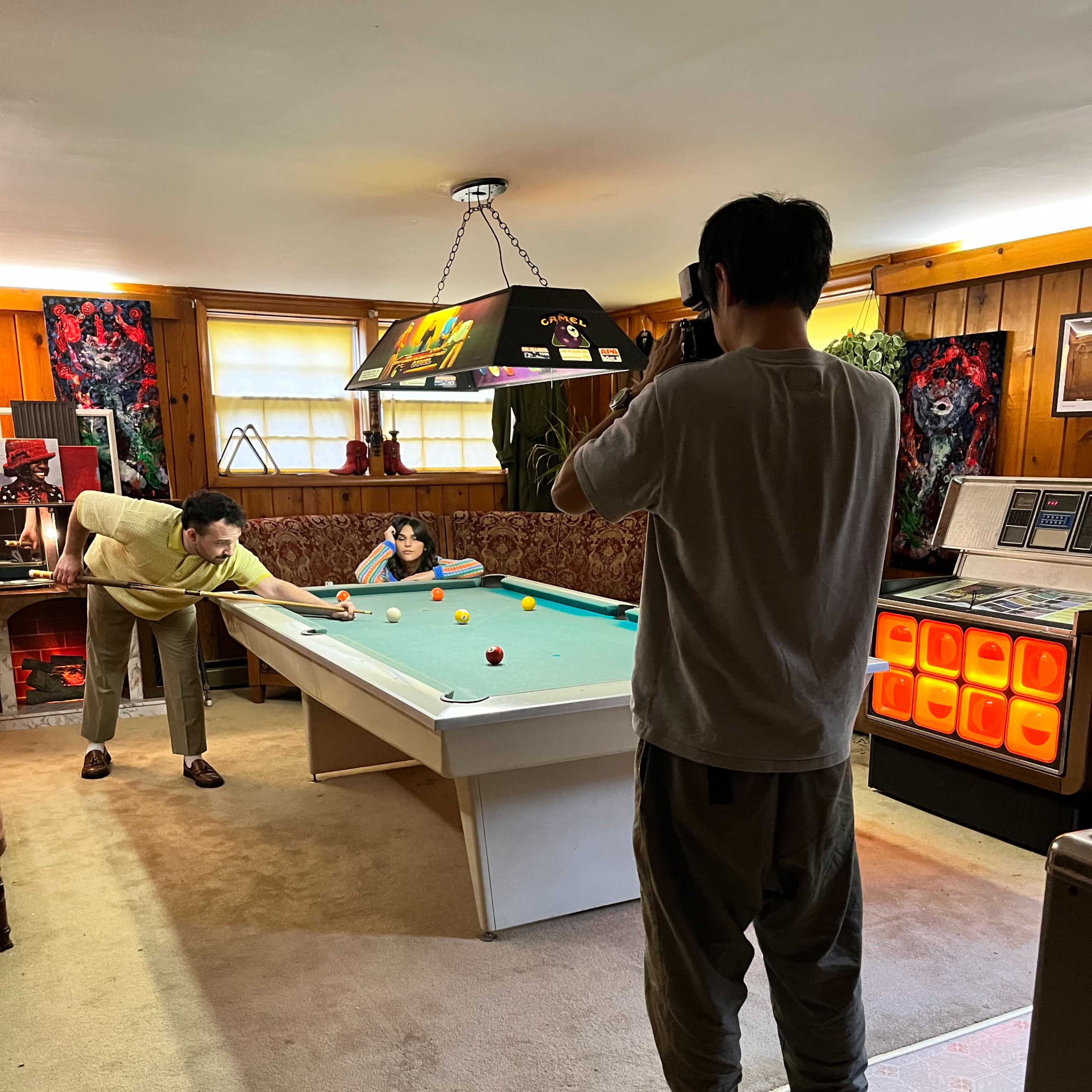 Two individuals are engaged in a billiards game in a room decorated with various artworks and a bright overhead light.