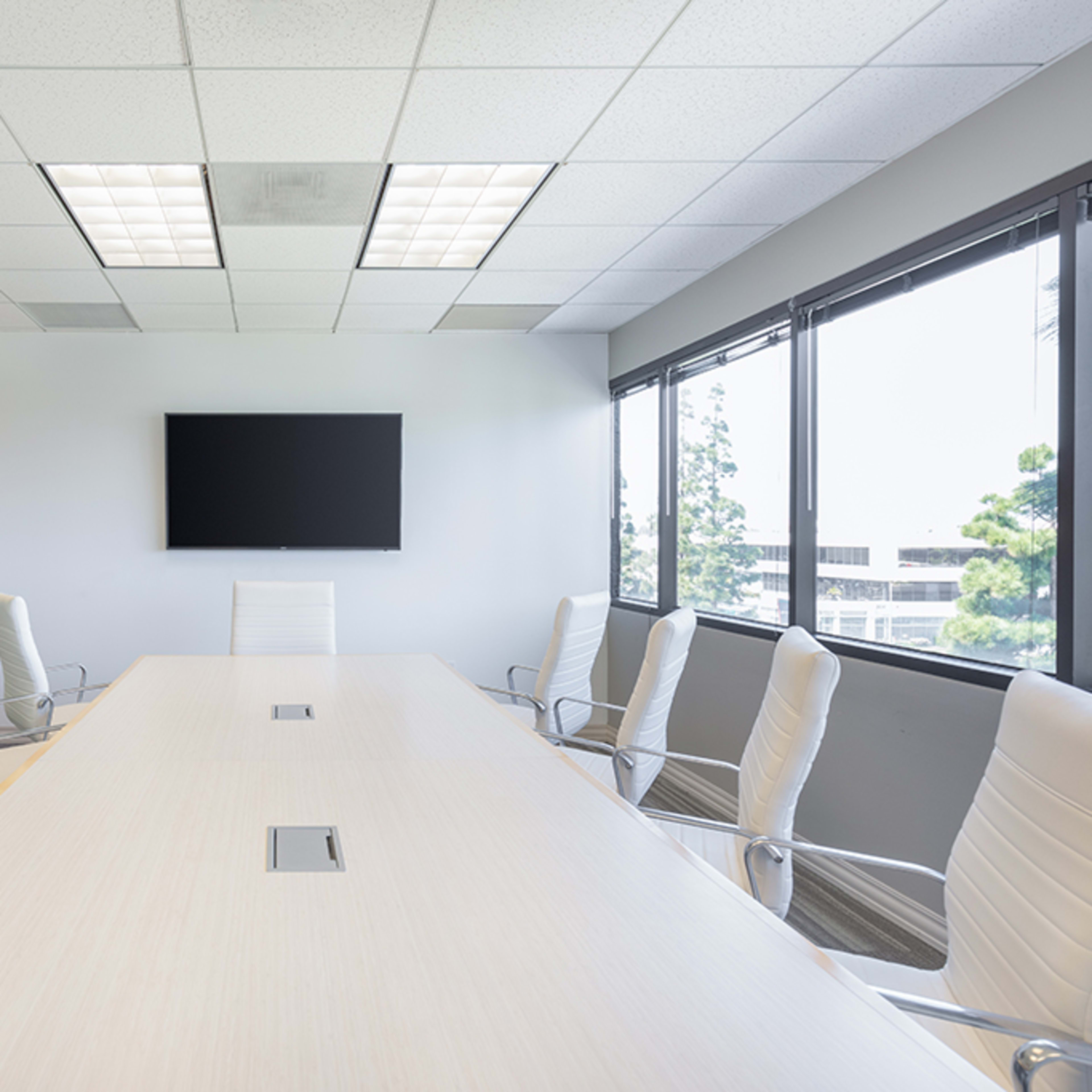 A conference room features a long table surrounded by white chairs, with large windows providing natural light and a wall-mounted television.