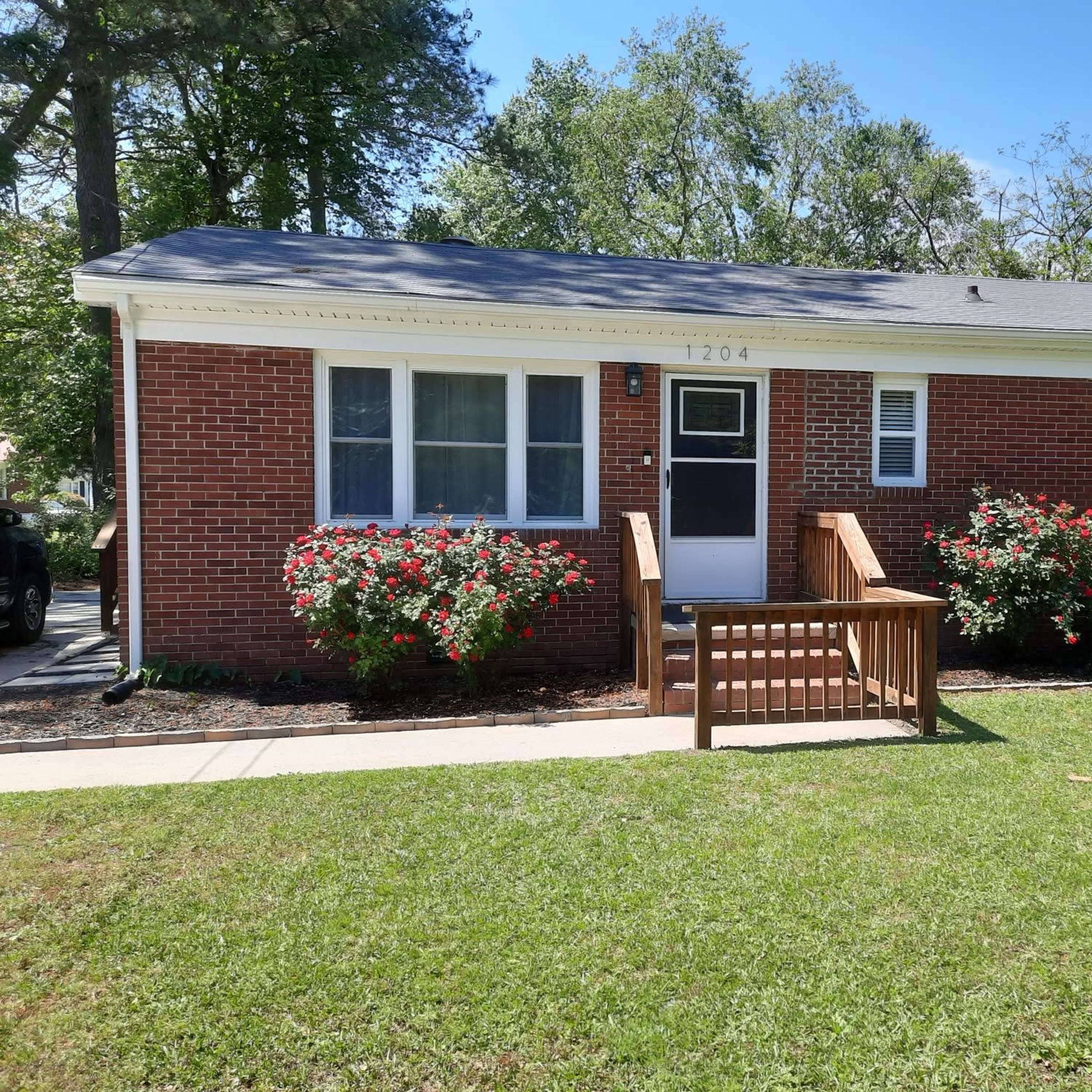A single-story brick house with a front wooden ramp and flower beds is set on a grassy lawn.