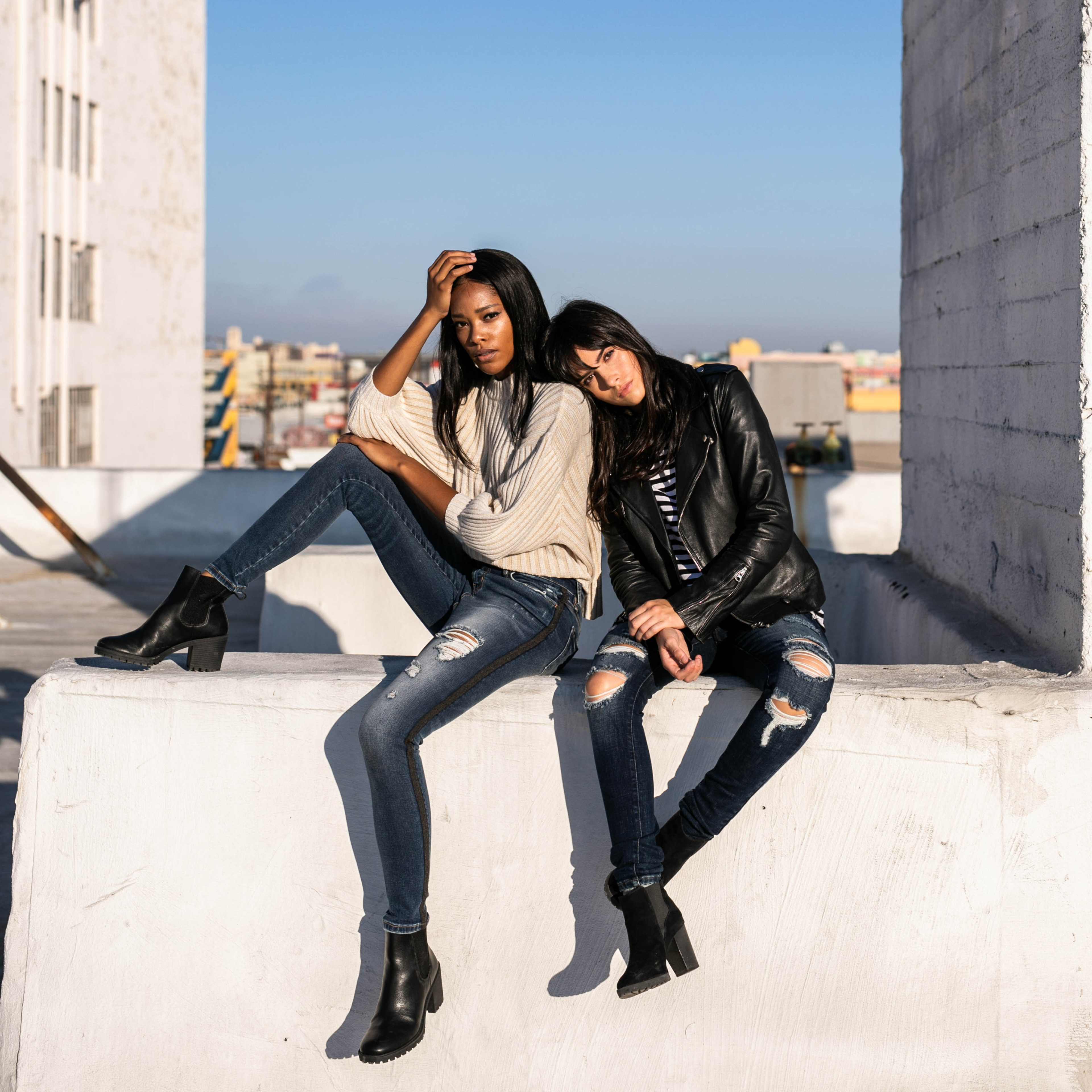 Two women sit on a concrete ledge wearing casual outfits, with one leaning back and the other resting her head on the first's shoulder, against a clear blue sky.