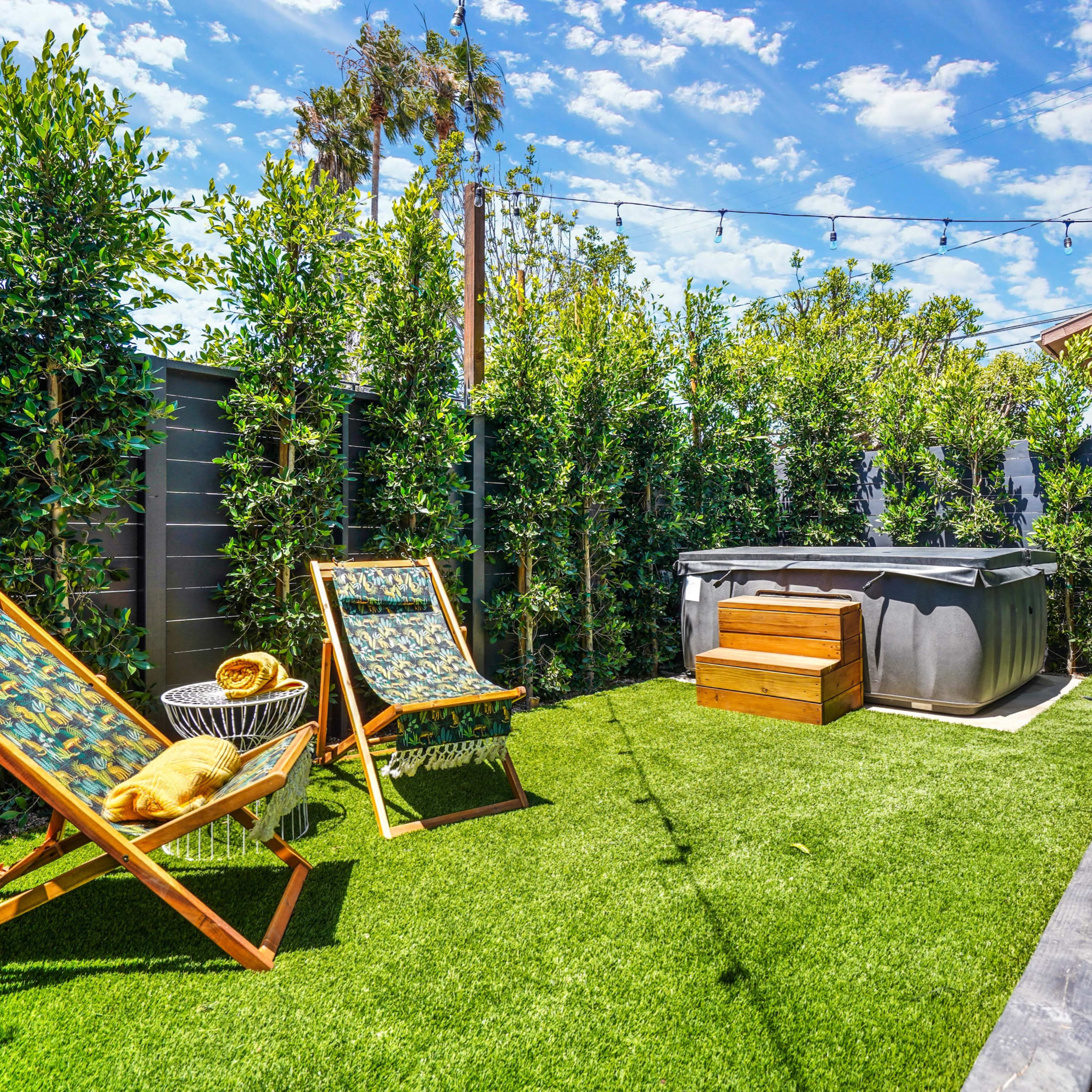 A backyard with two lounge chairs, a hot tub, and lush greenery under a blue sky with scattered clouds.