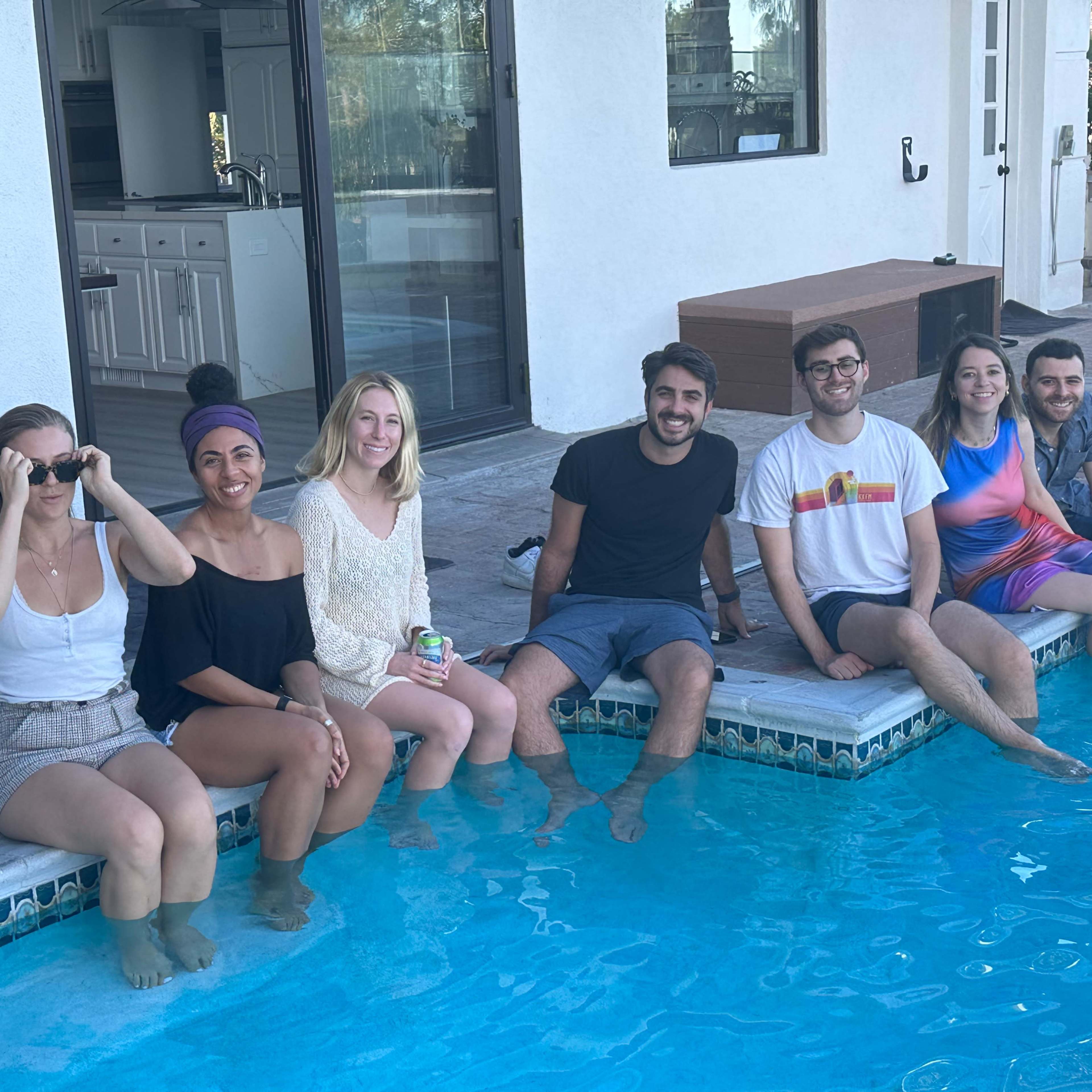 A group of seven people sits on the edge of a swimming pool, dipping their feet in the water, with a house visible in the background.