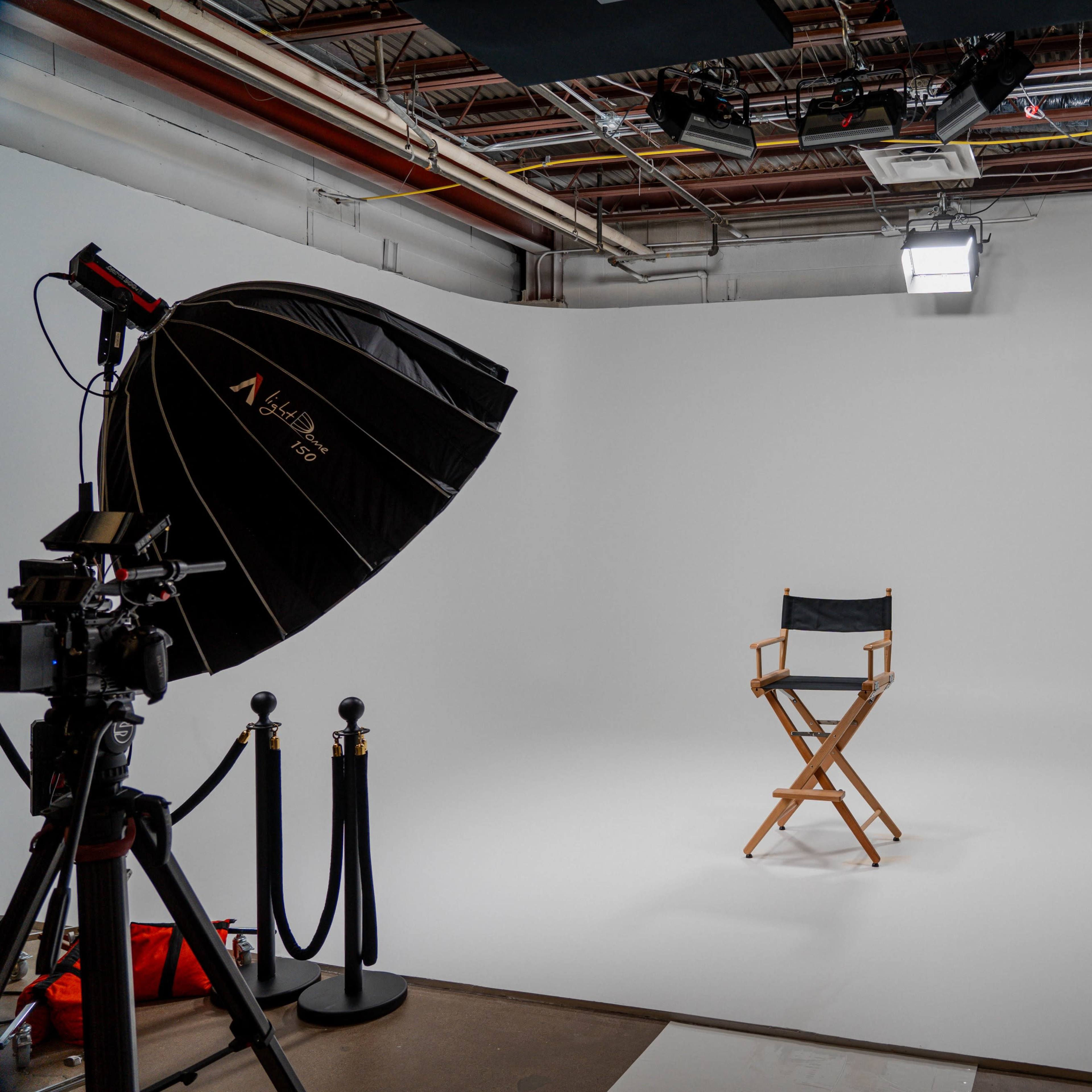 A photography studio features a black director's chair centered in front of a white backdrop, with lighting equipment and a camera positioned nearby.