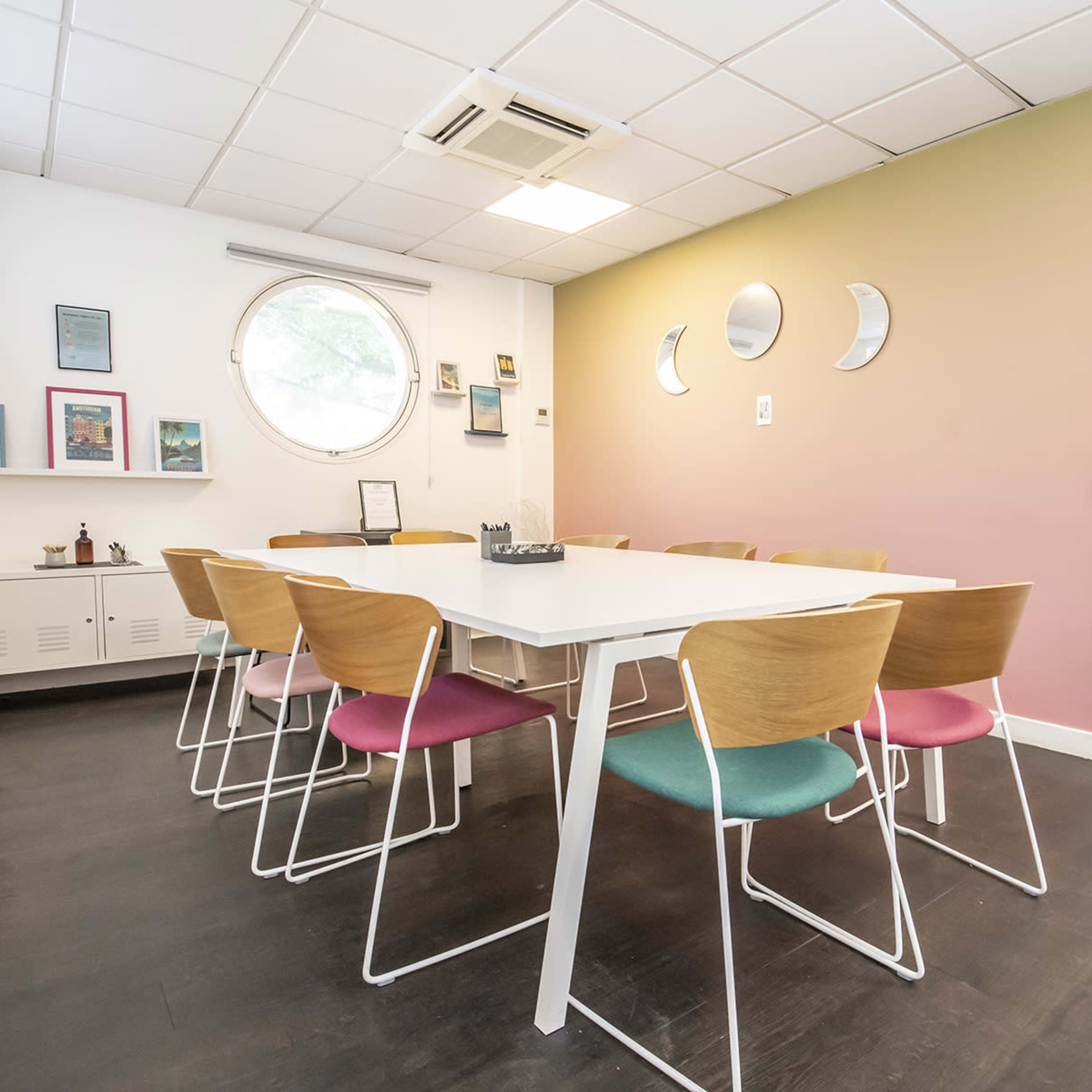 A minimalist meeting room features a white table surrounded by wooden chairs with colored cushions, positioned against a partially painted wall with circular mirrors.