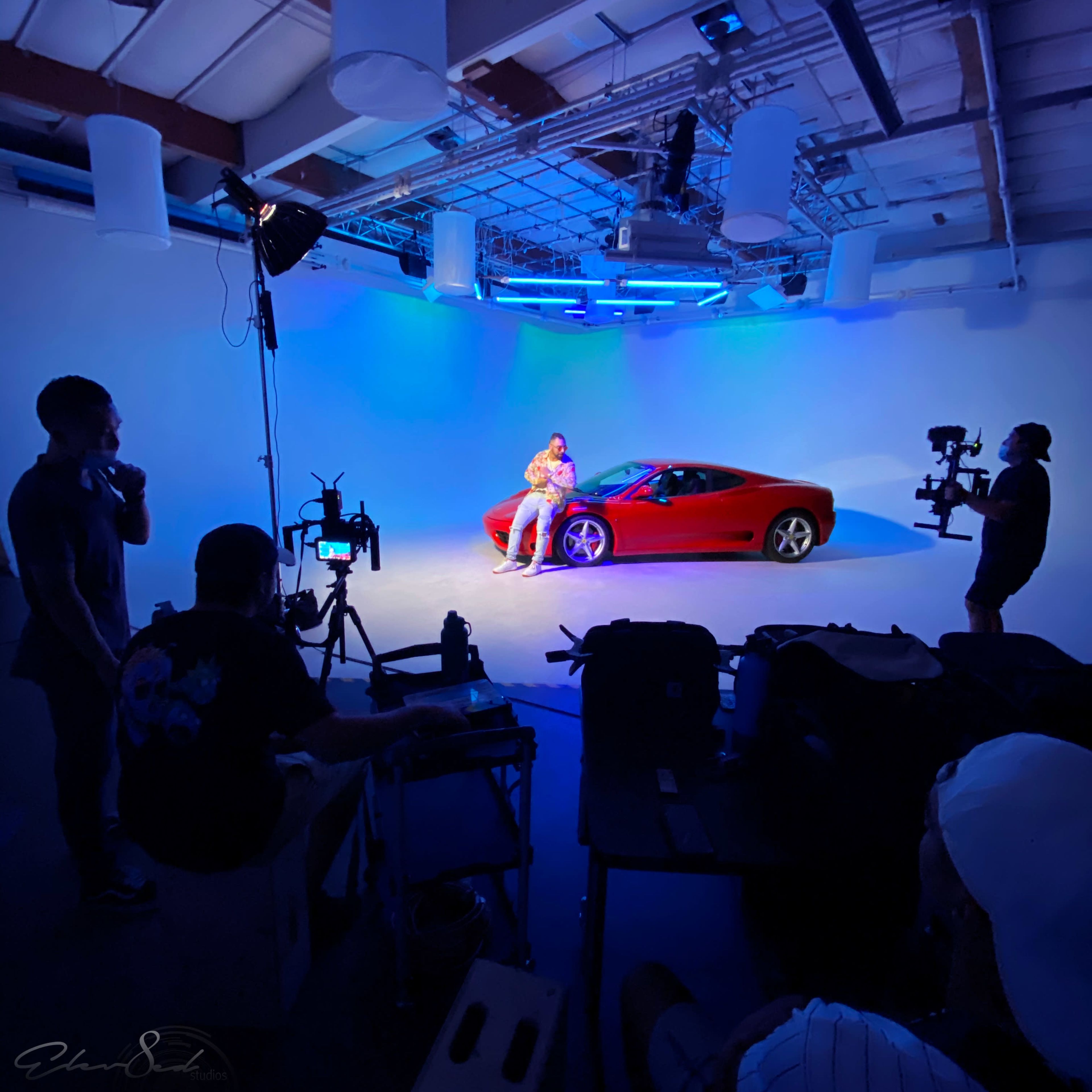 A red sports car is positioned in a studio with blue lighting, while a person sits on the car and a small crew operates cameras and equipment nearby.