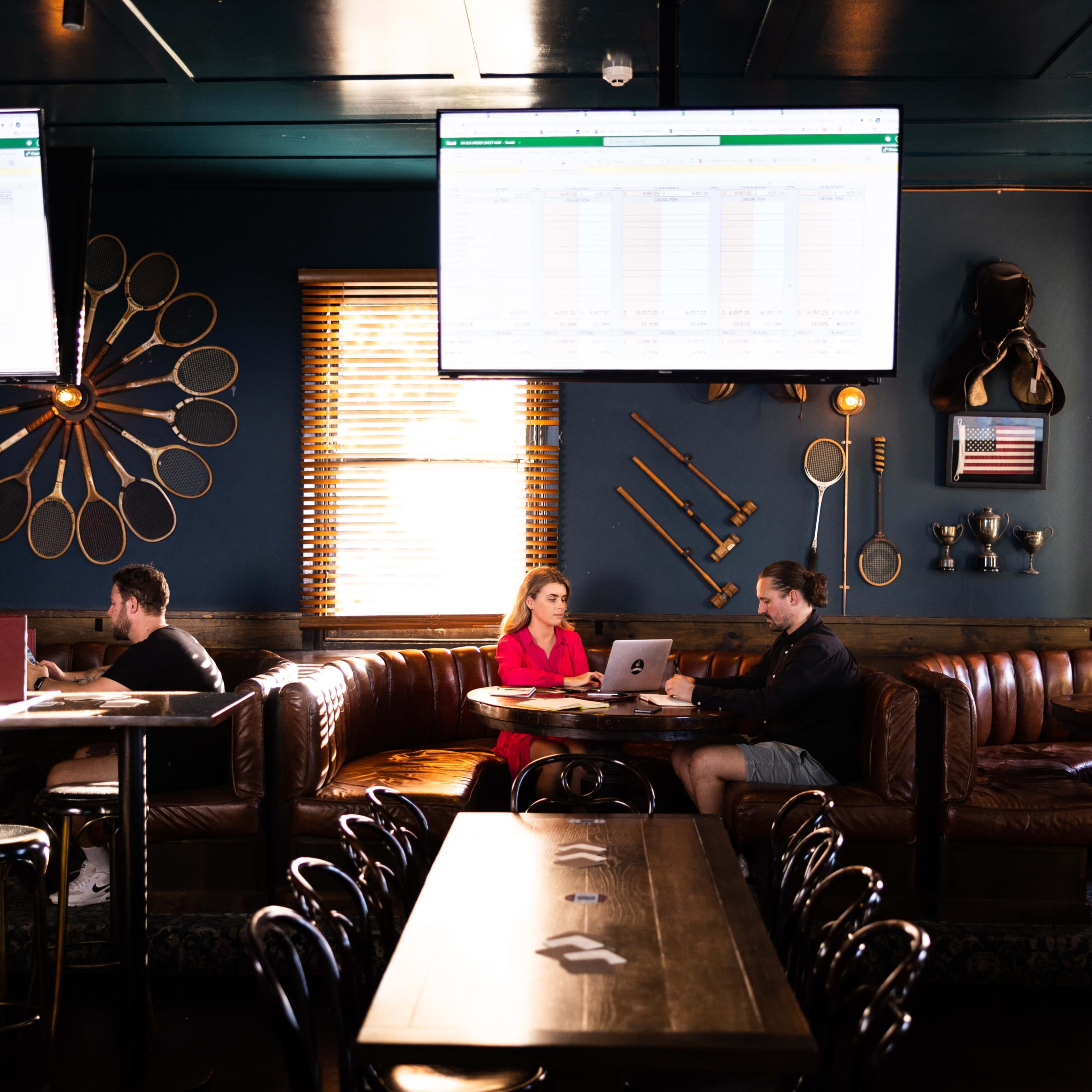 A restaurant interior features dark walls, leather seating, and large screens displaying data, with three patrons engaged in work and conversation.