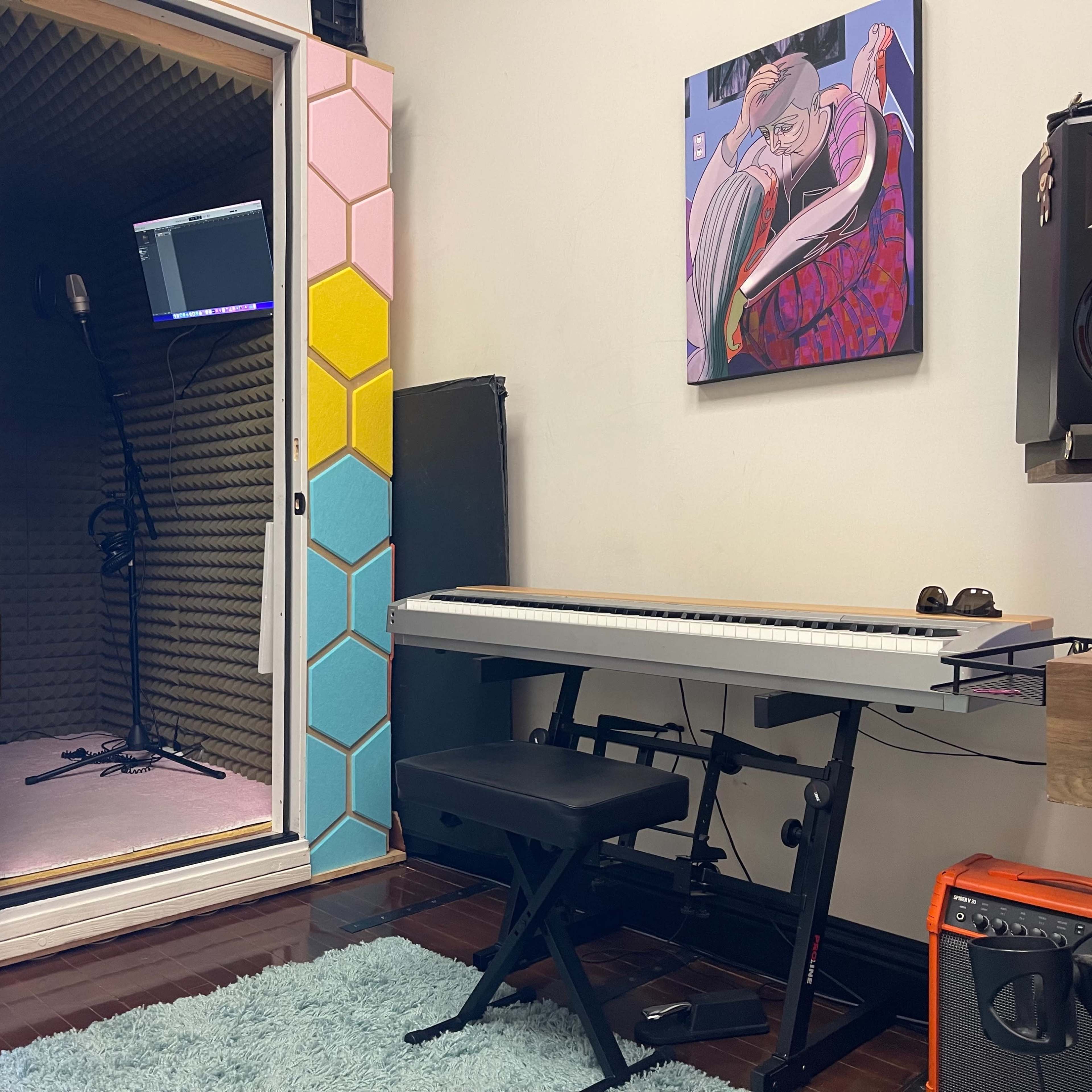 A music studio features a soundproof booth with a microphone, a keyboard on a stand, and an amplifier beside a colorful artwork on the wall.