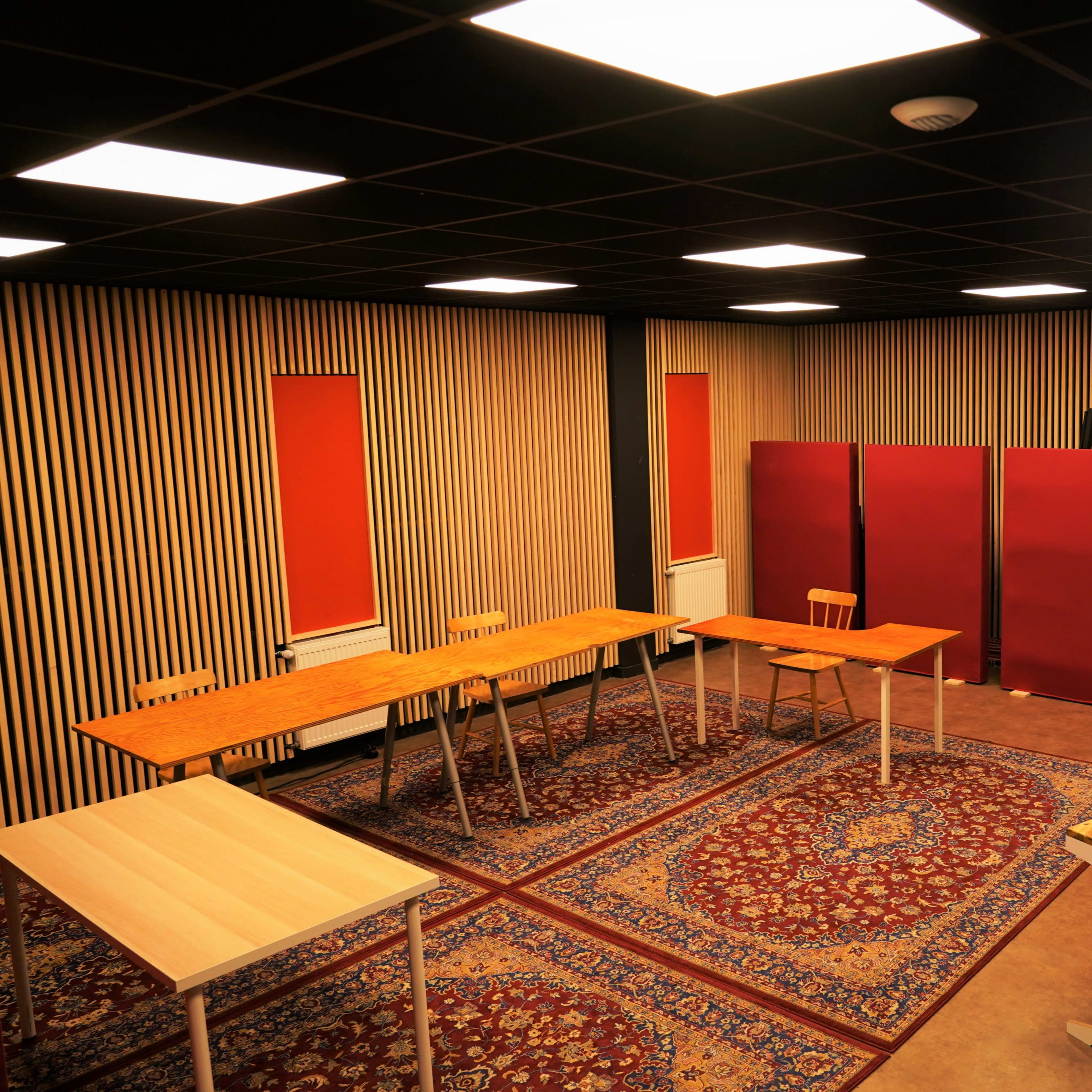 A room with several wooden tables arranged on a patterned rug, flanked by red window coverings and divided by red partitions.