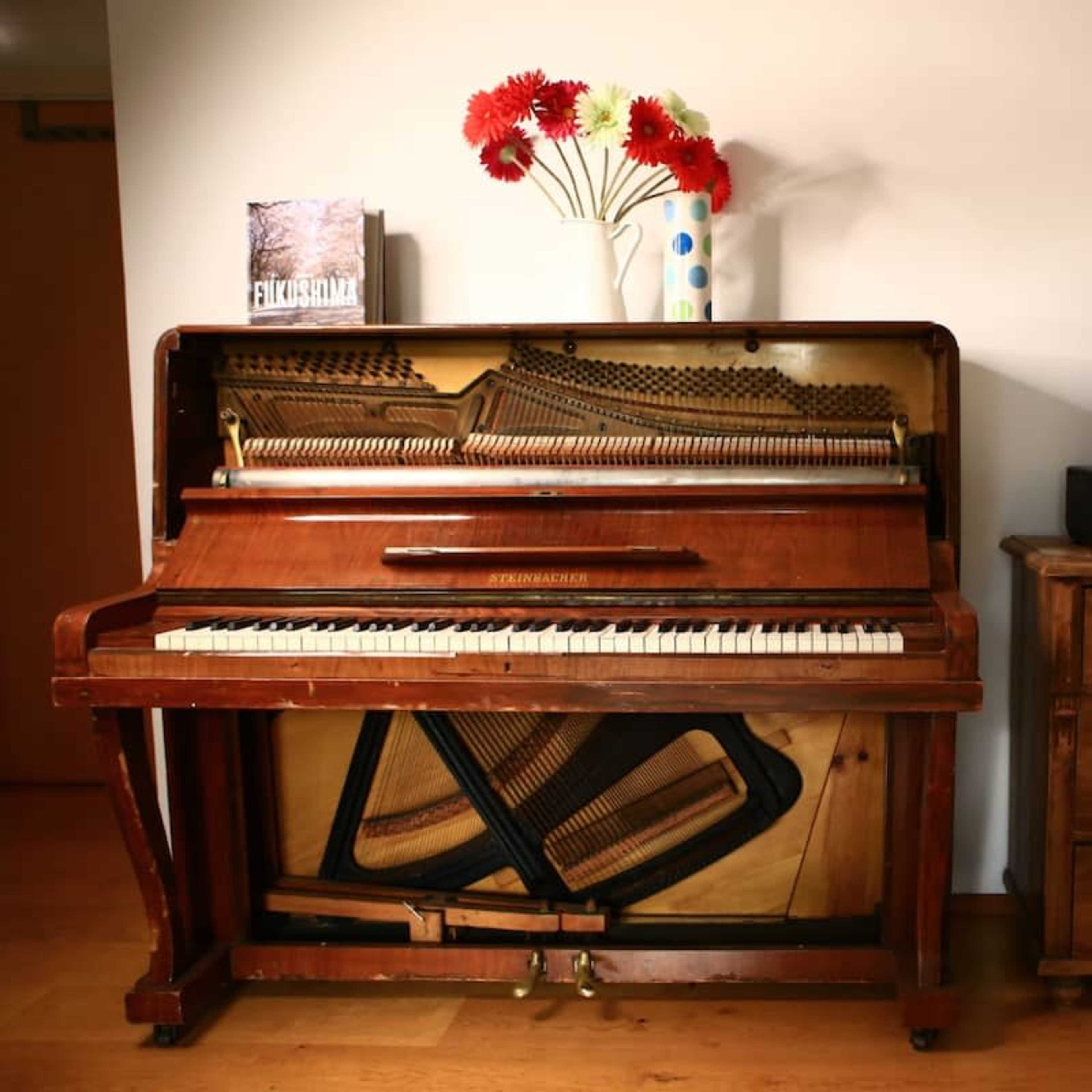 A vintage upright piano stands next to a wooden dresser, adorned with a vase of flowers and a small decorative item.