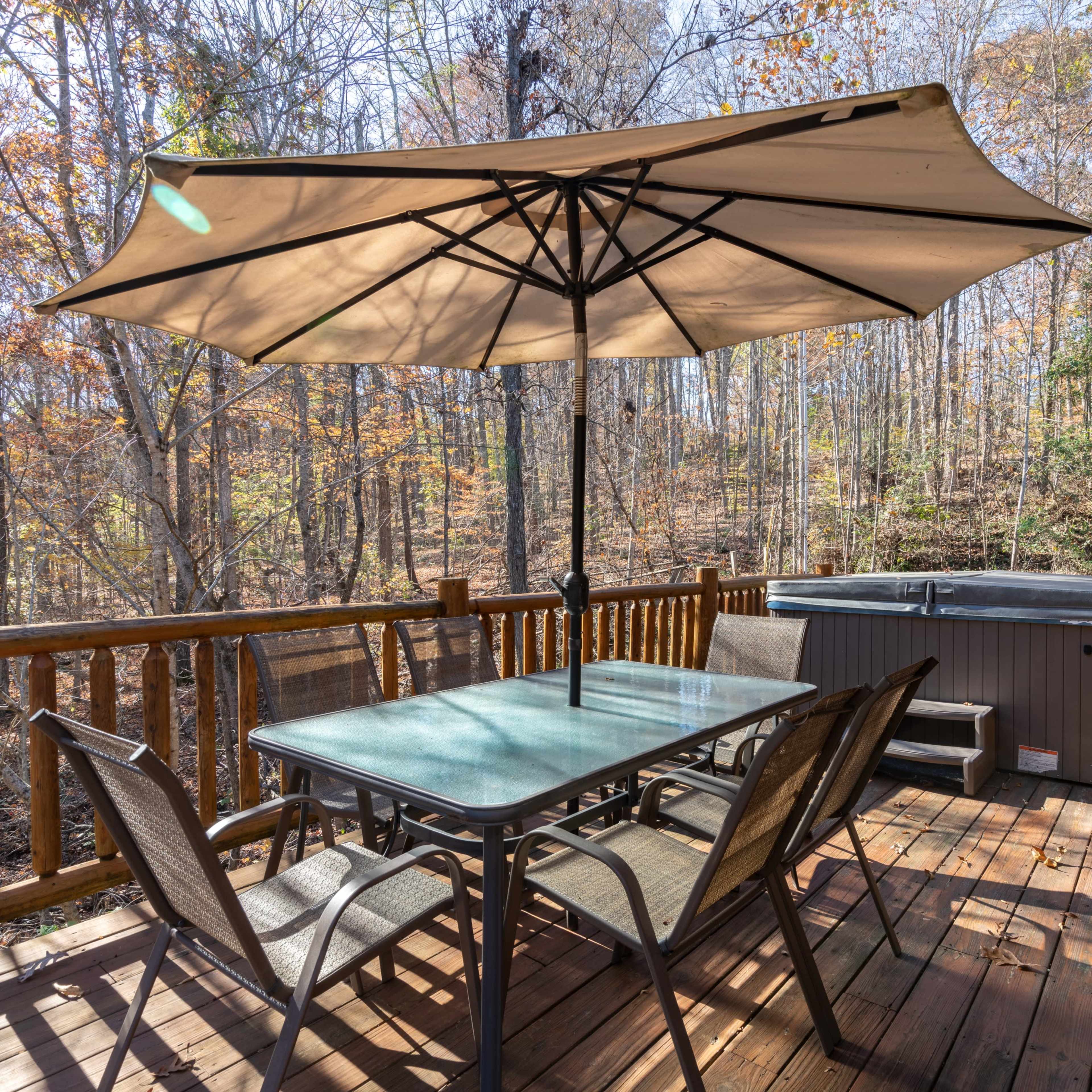 A deck features a large umbrella over a glass table surrounded by chairs, with a hot tub and wooded area in the background.
