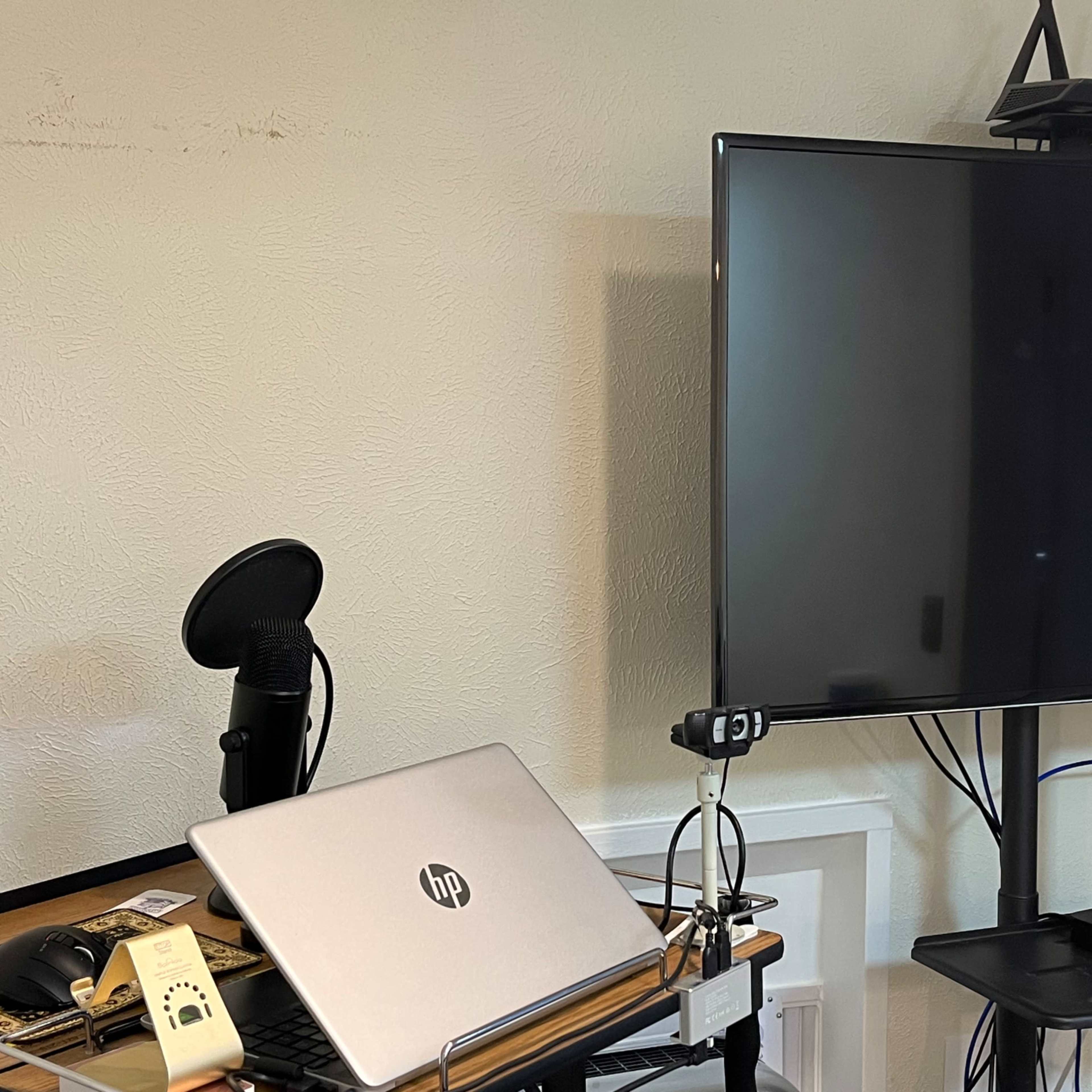 A silver HP laptop sits on a desk beside a microphone and a large wall-mounted television.