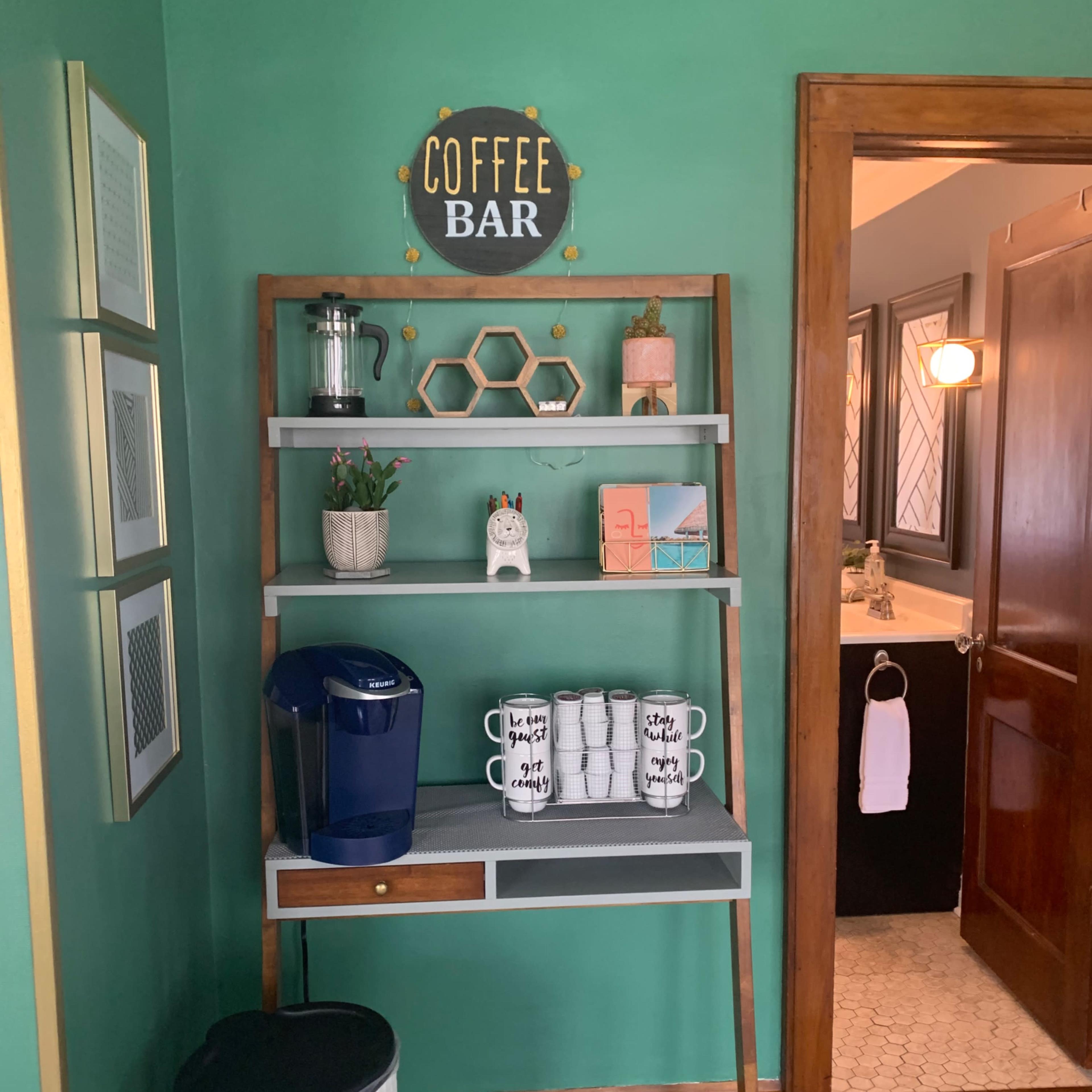 A wooden shelving unit displaying coffee supplies and decorative items against a green wall, with a sign that reads "COFFEE BAR."
