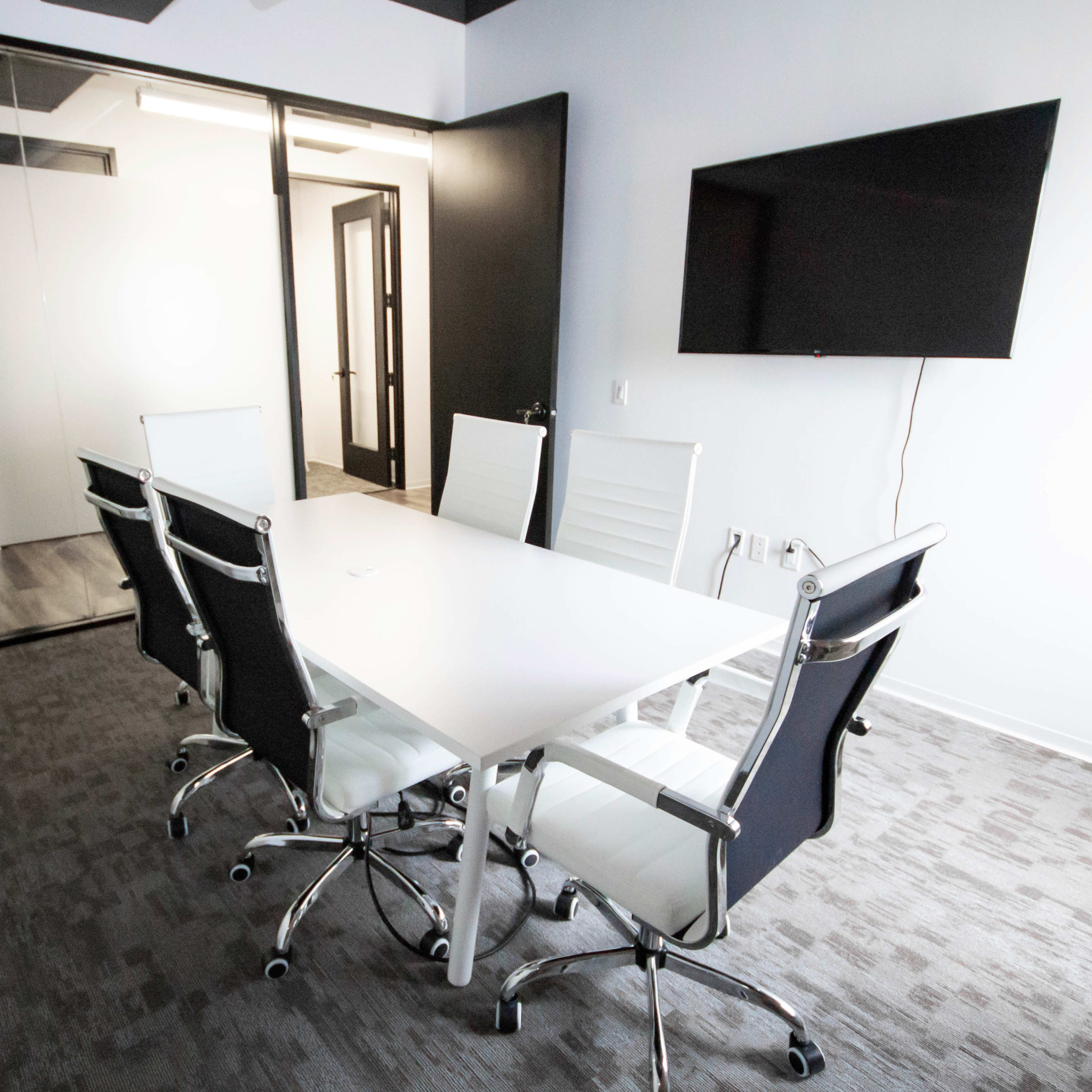 A modern conference room with a white table and six chairs, a large screen mounted on the wall, and a glass wall separating it from an adjacent space.