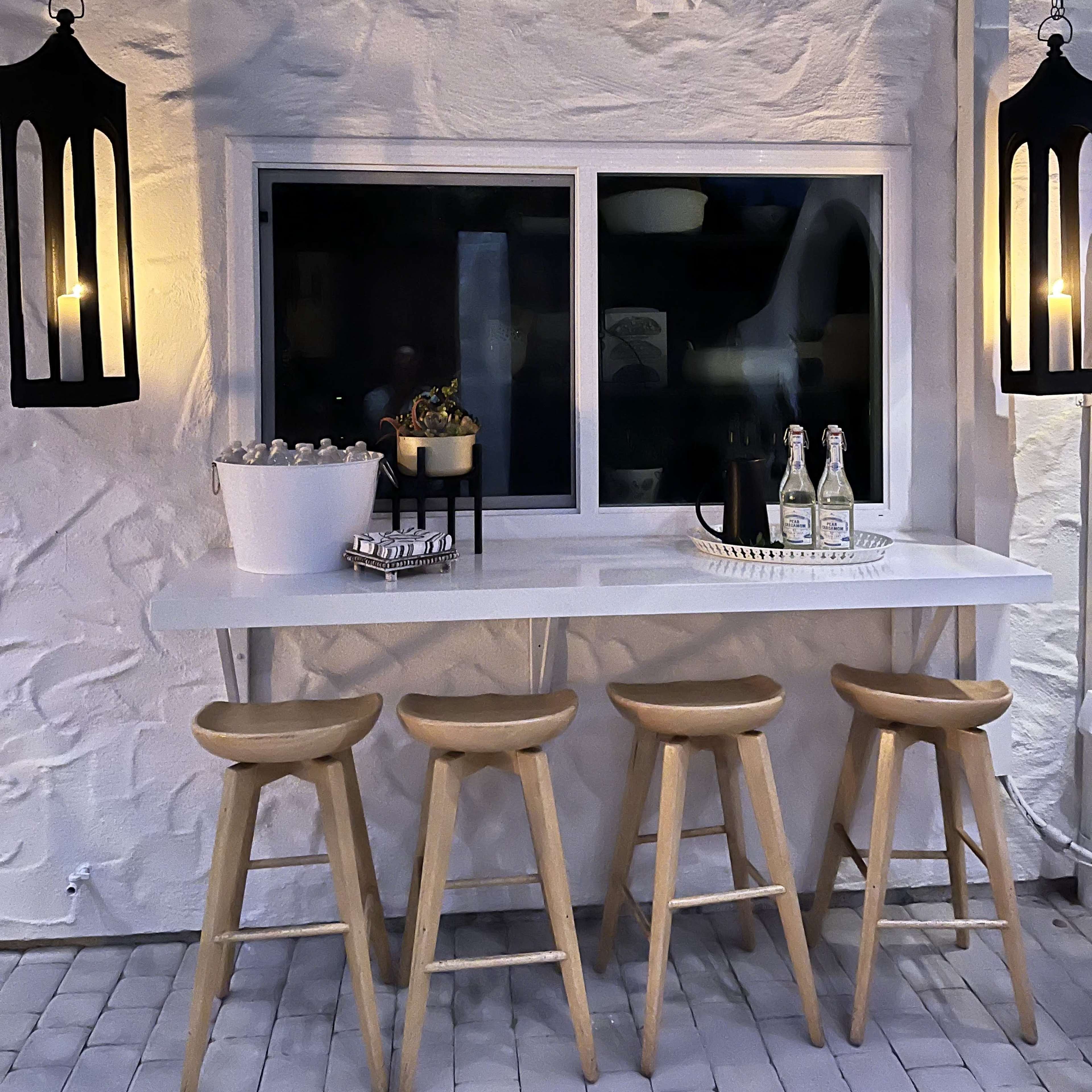 A minimalist bar area features four wooden stools and a countertop with a white backdrop and illuminated lanterns hanging on either side.