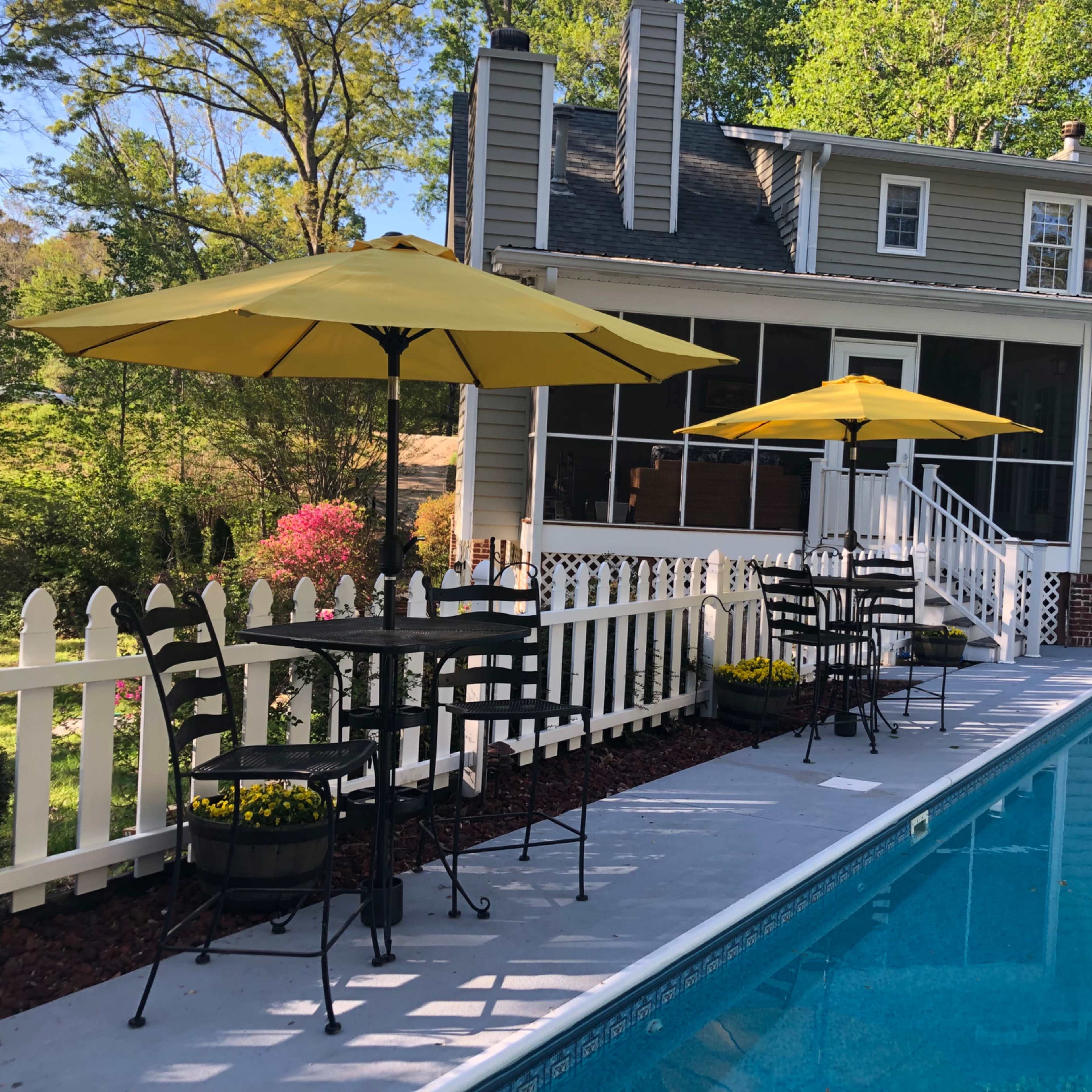 A backyard pool area features two yellow umbrellas over black metal tables beside the water, with a fenced garden in the background.