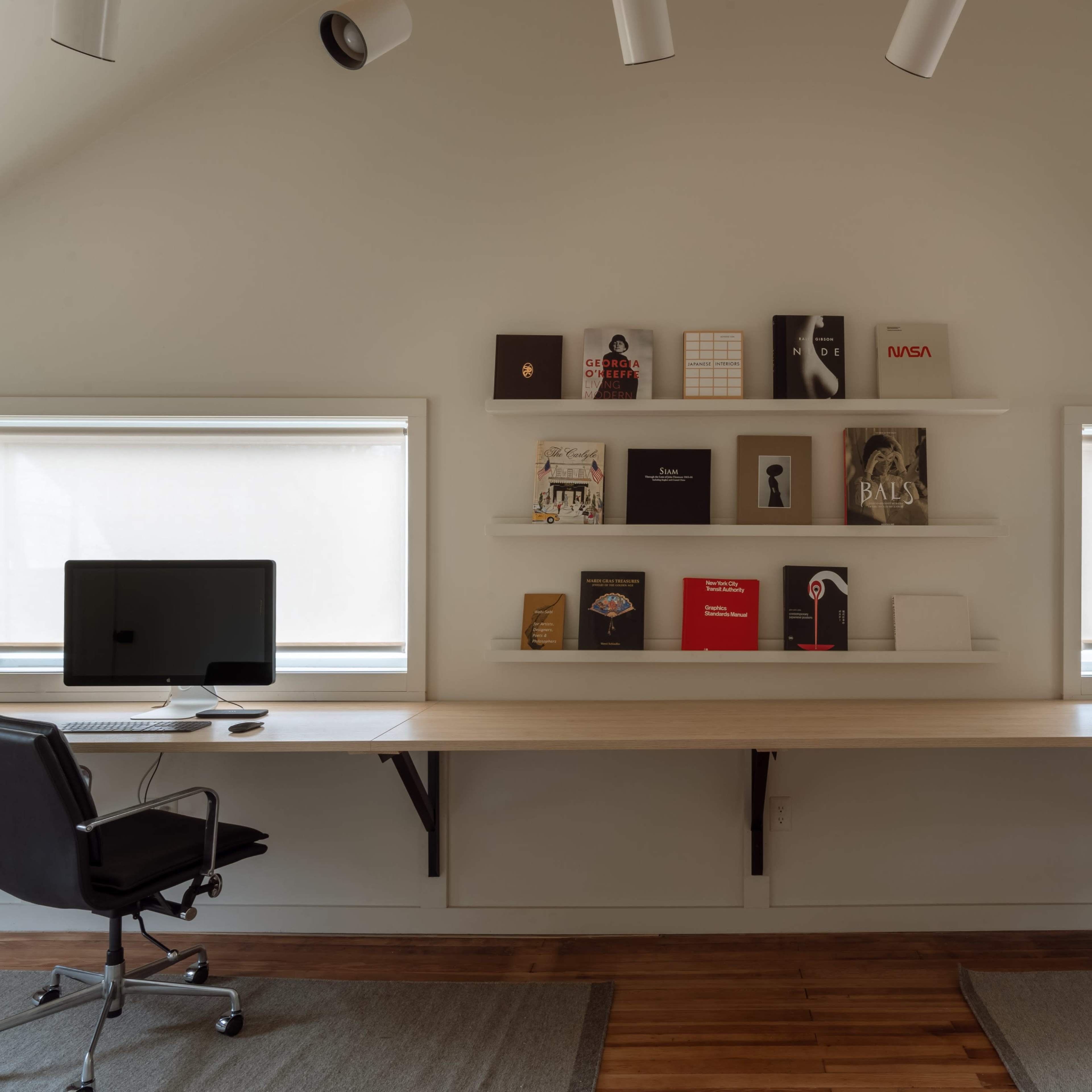 A modern workspace with two computers on a long wooden desk, flanked by shelves displaying various books and magazines above, along with two large windows.