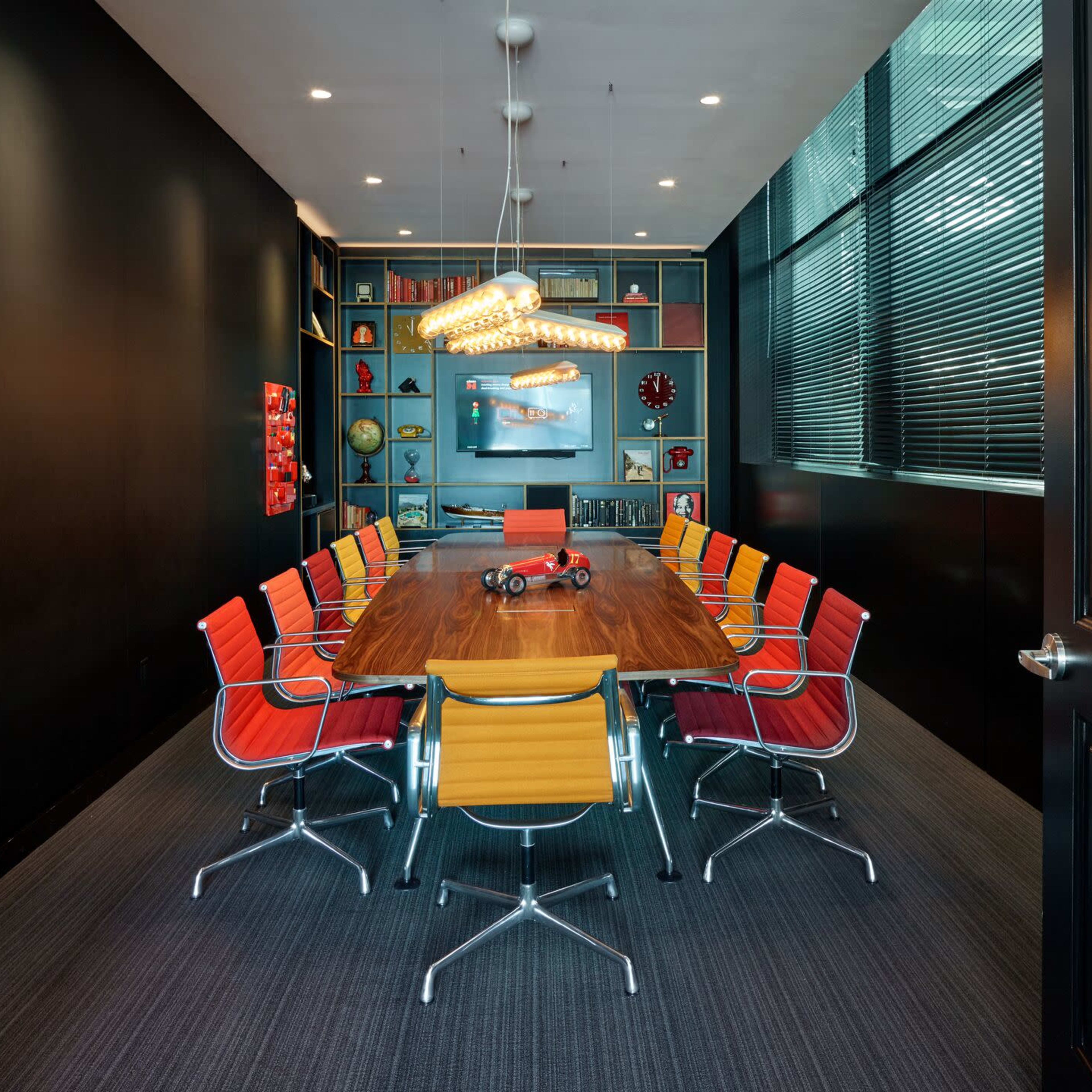 A modern conference room features a long wooden table surrounded by colorful chairs, with shelves displaying decorative items in the background.