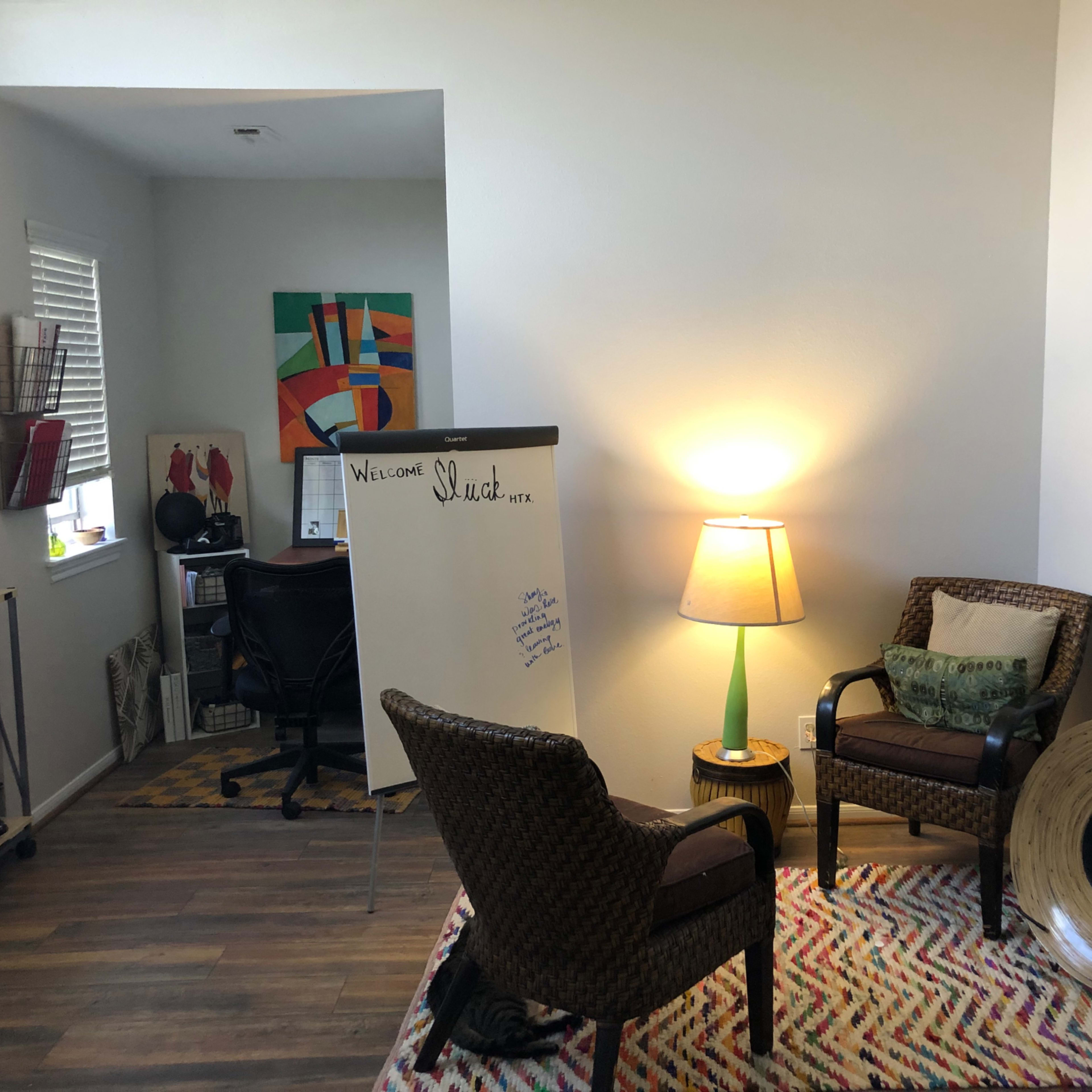 A well-lit room featuring two woven chairs, a lamp, a whiteboard, and a desk area with artwork on the walls.