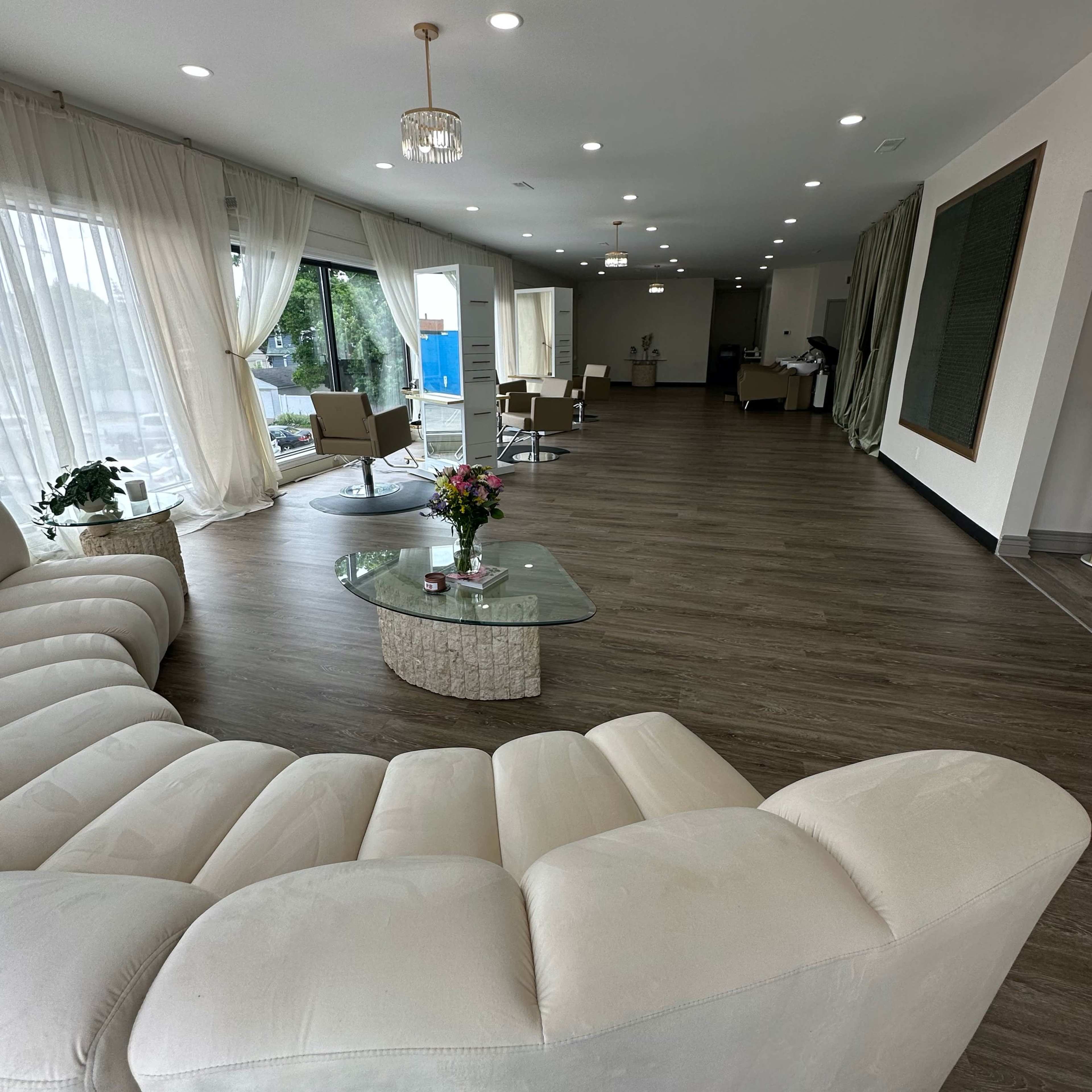 A spacious, modern interior featuring a light-colored sofa, a glass coffee table, and large windows draped with sheer curtains.