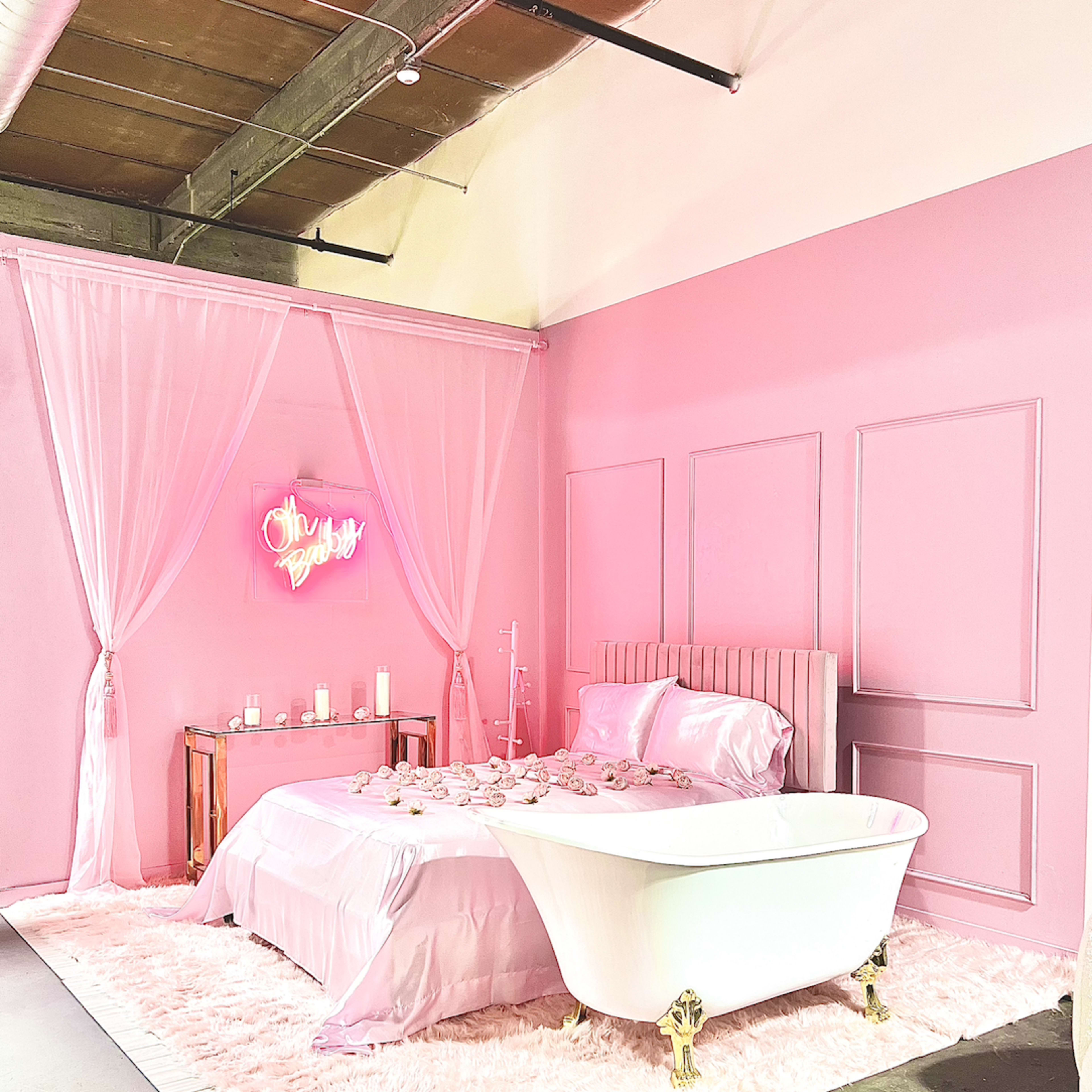 The scene features a pink-themed bedroom with a clawfoot bathtub, a bed adorned with silky linens, and decorative elements like candles on a side table.