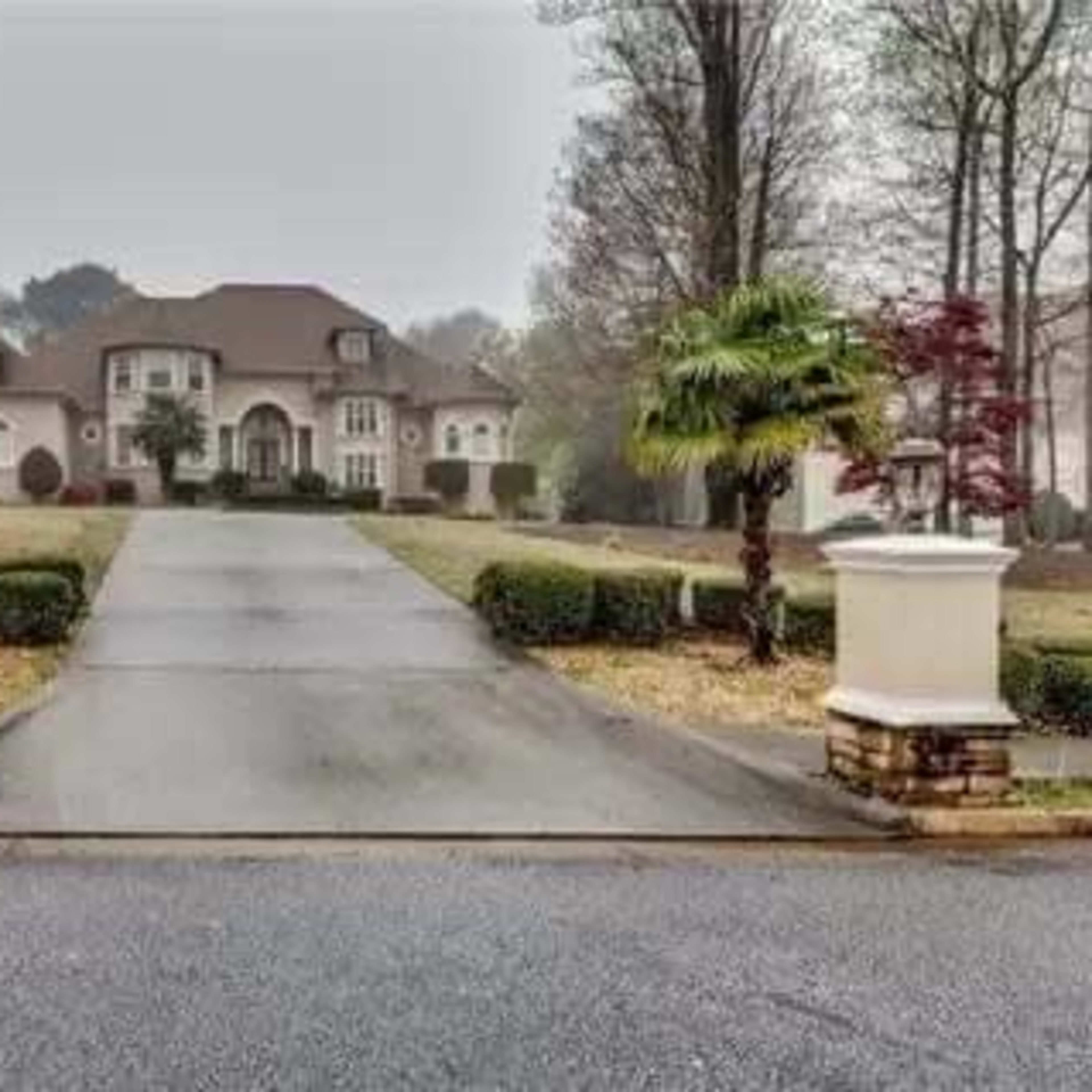 A large, upscale house set back from the road with a long driveway, flanked by palm trees and landscaped greenery.