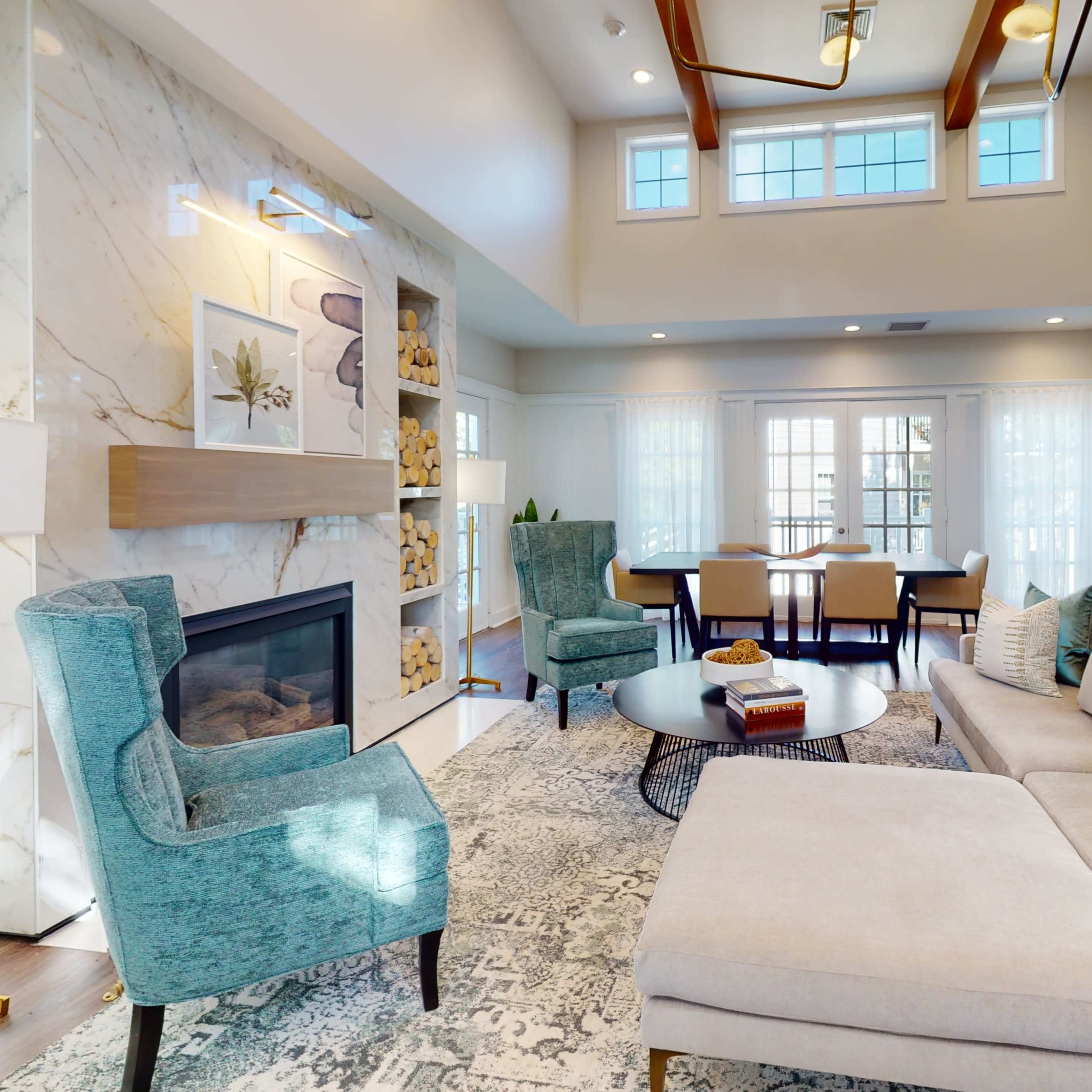 A modern living room with a sectional sofa, two blue chairs, a round coffee table, and a fireplace, all under a high ceiling with large windows allowing natural light.