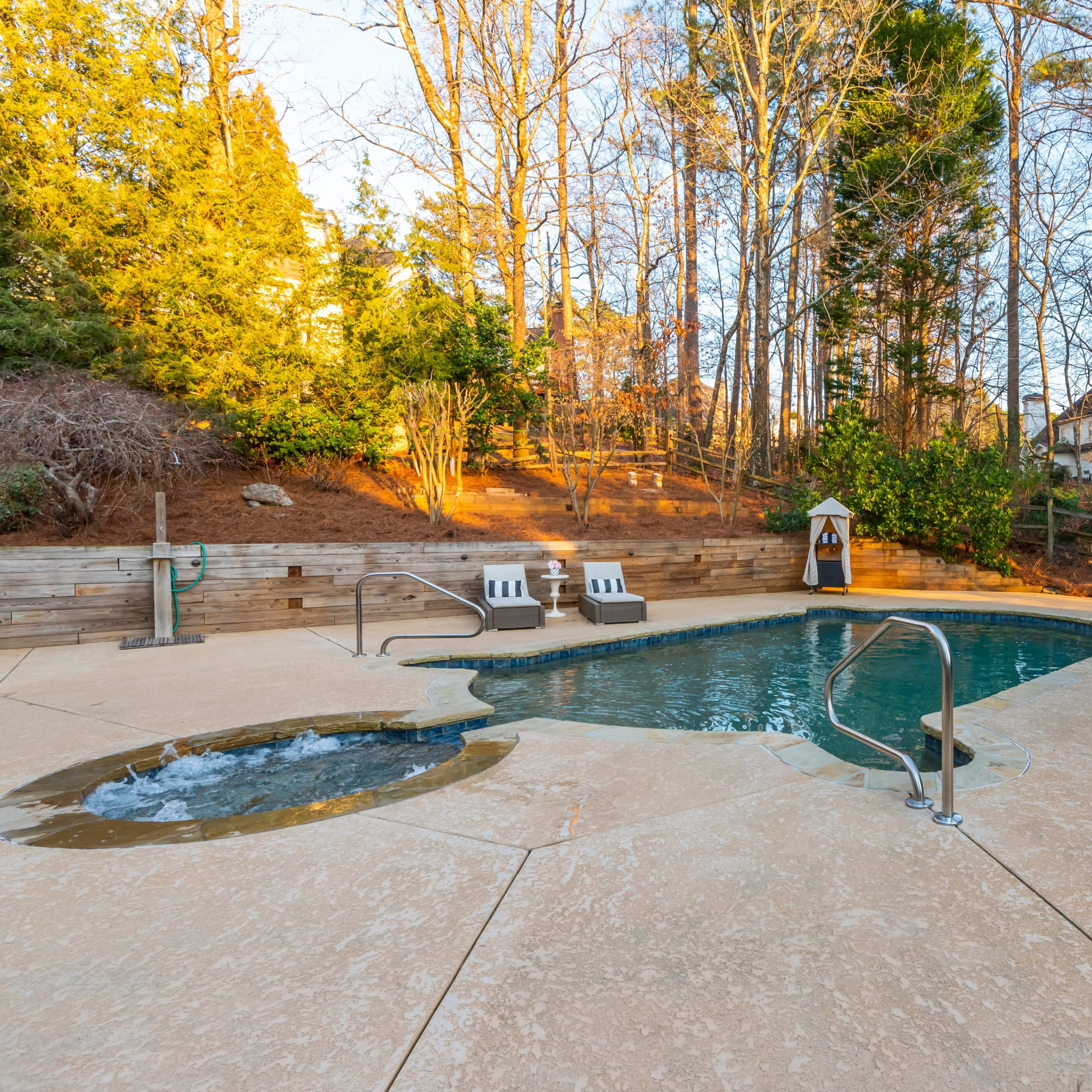 A rectangular swimming pool with a spa is surrounded by a stone deck and a gazebo, set against a backdrop of trees and a residential area.