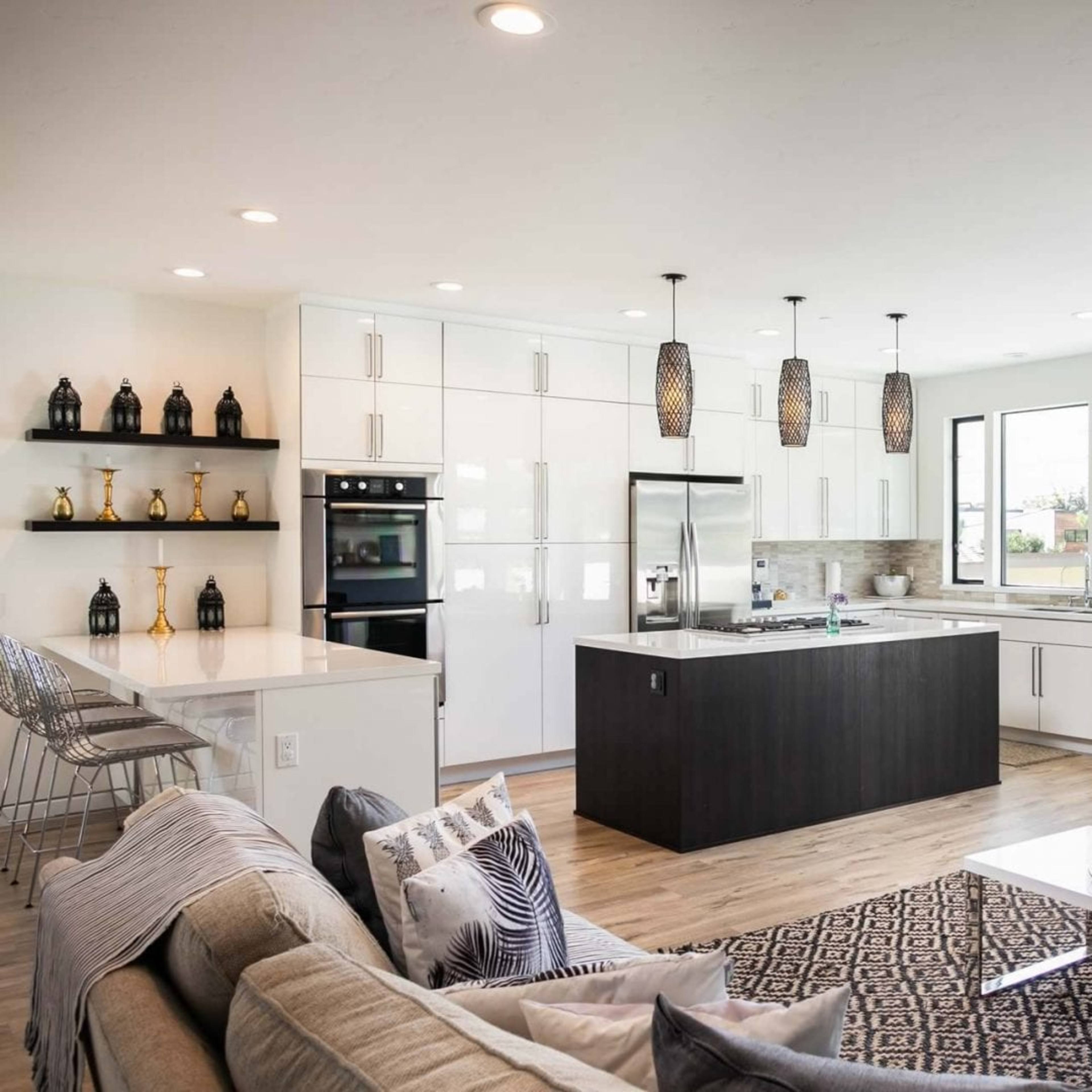 A modern kitchen and living area with white cabinetry, a central island, and large windows allowing natural light to fill the space.