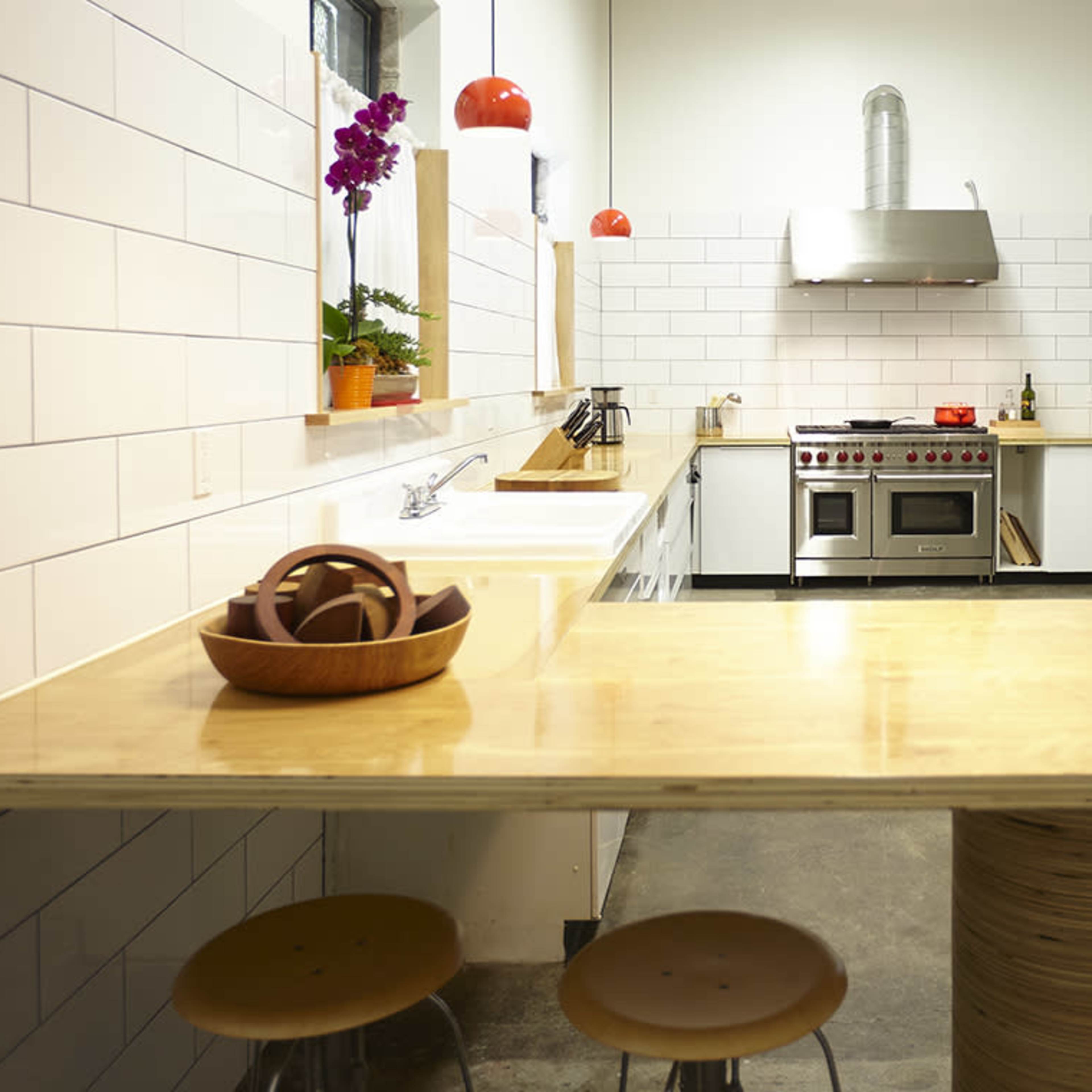 A modern kitchen with white tiled walls, a central wooden table, and stainless steel appliances, including a large oven and refrigerator.