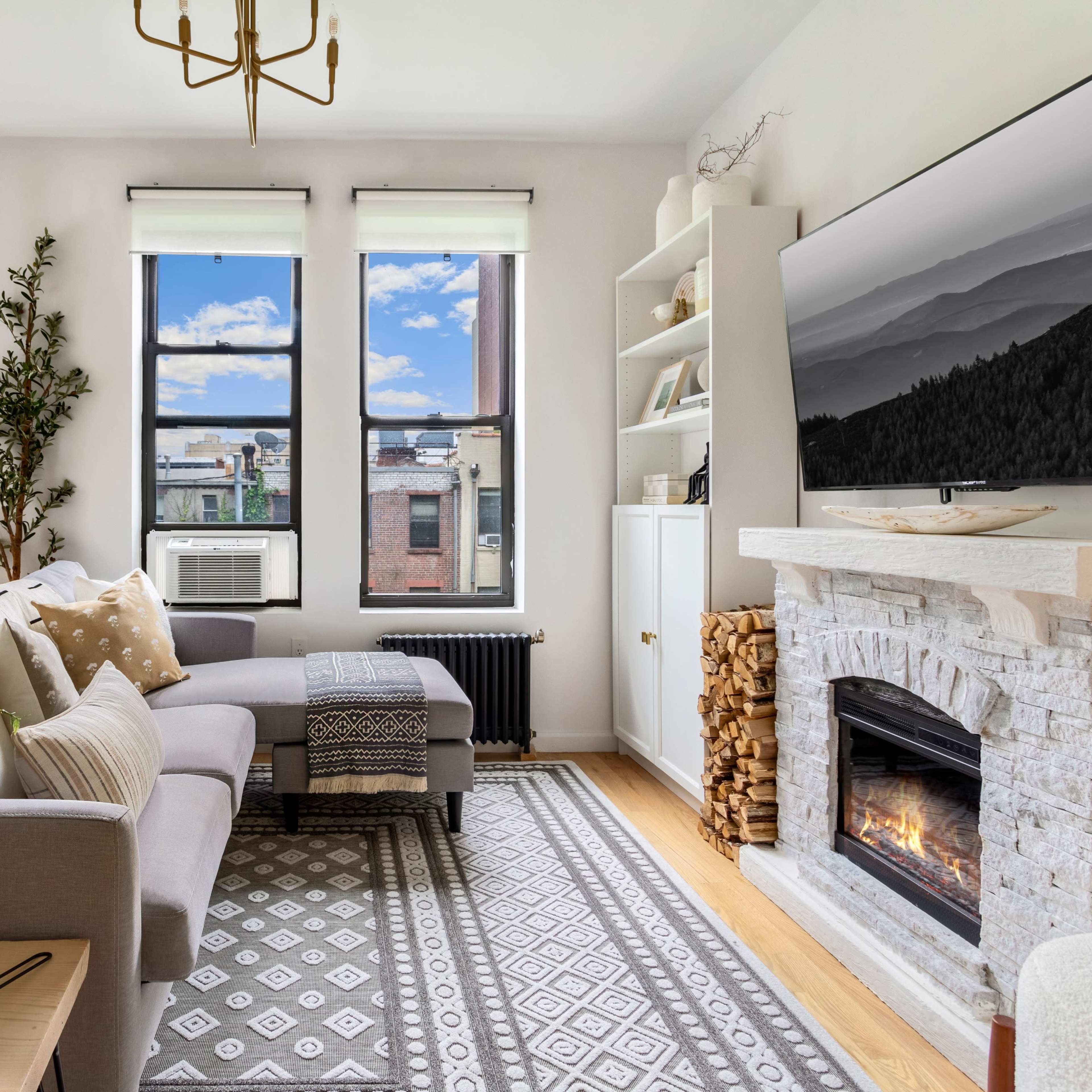 A cozy living room features a sectional sofa, large windows with views of urban scenery, a decorative stone fireplace, and shelves filled with plants and decor.