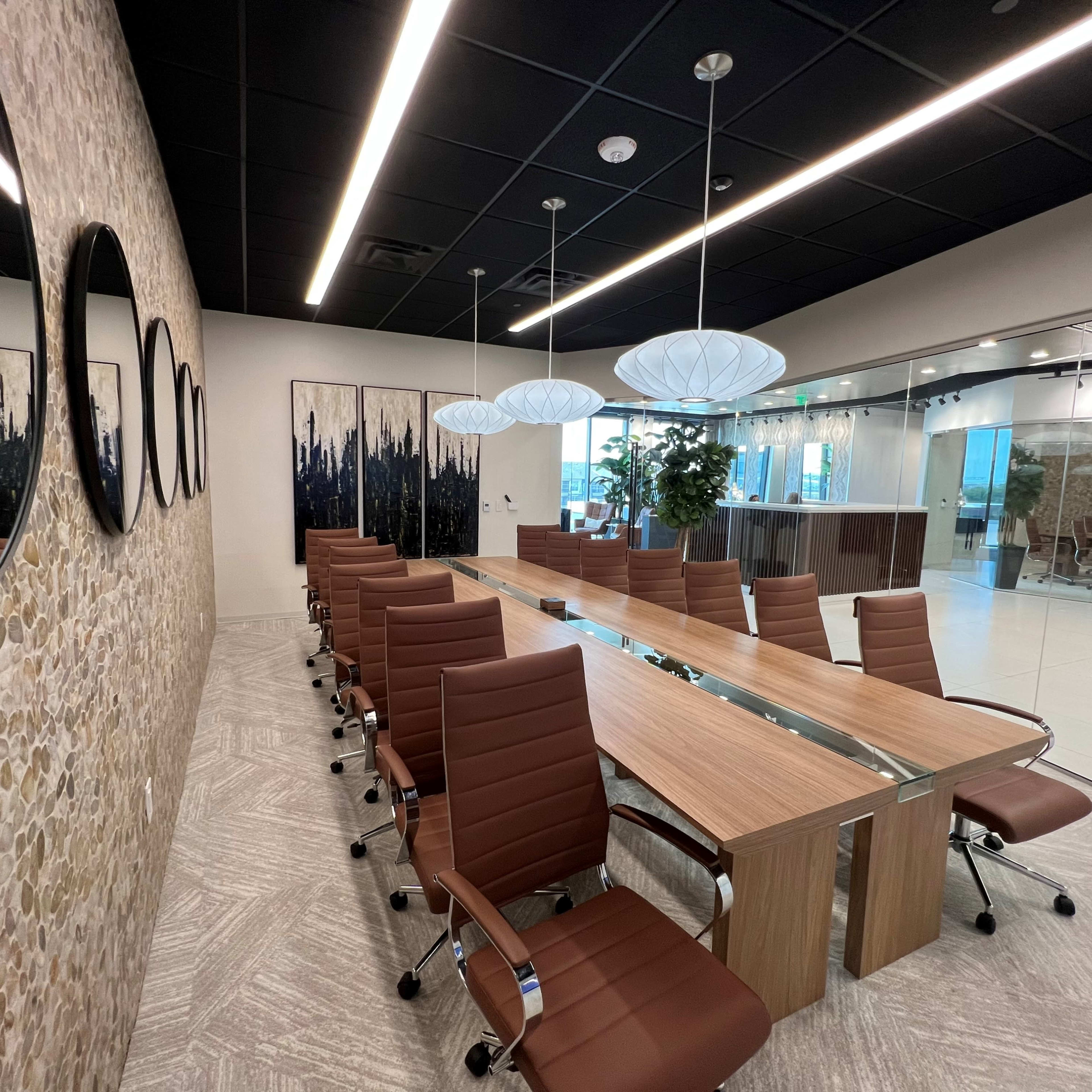 A modern conference room features a long wooden table surrounded by brown leather chairs, with stone-textured walls and large mirrors, complemented by artistic wall panels and overhead lighting.