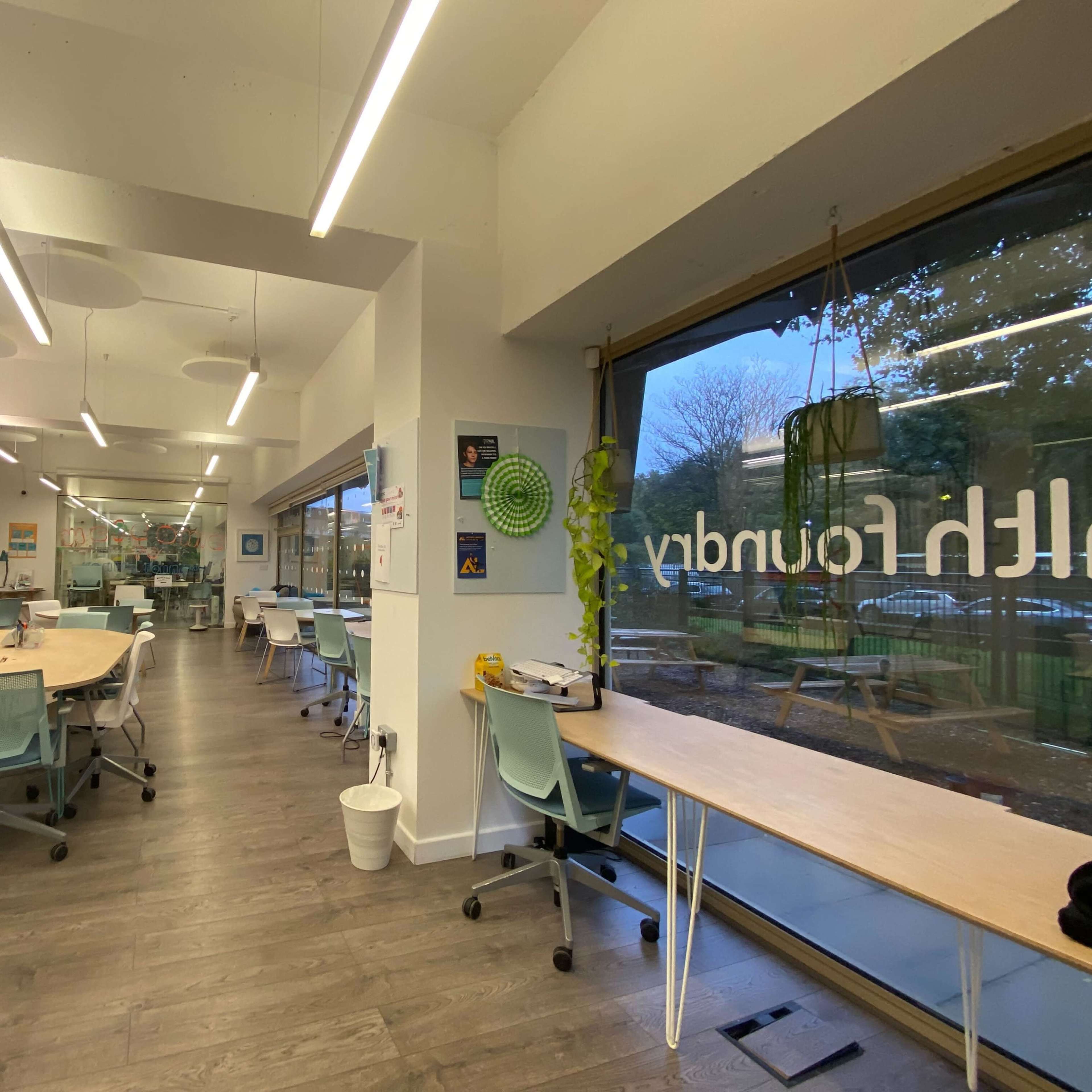 A modern, open workspace with several tables and chairs arranged in a well-lit room featuring large windows.
