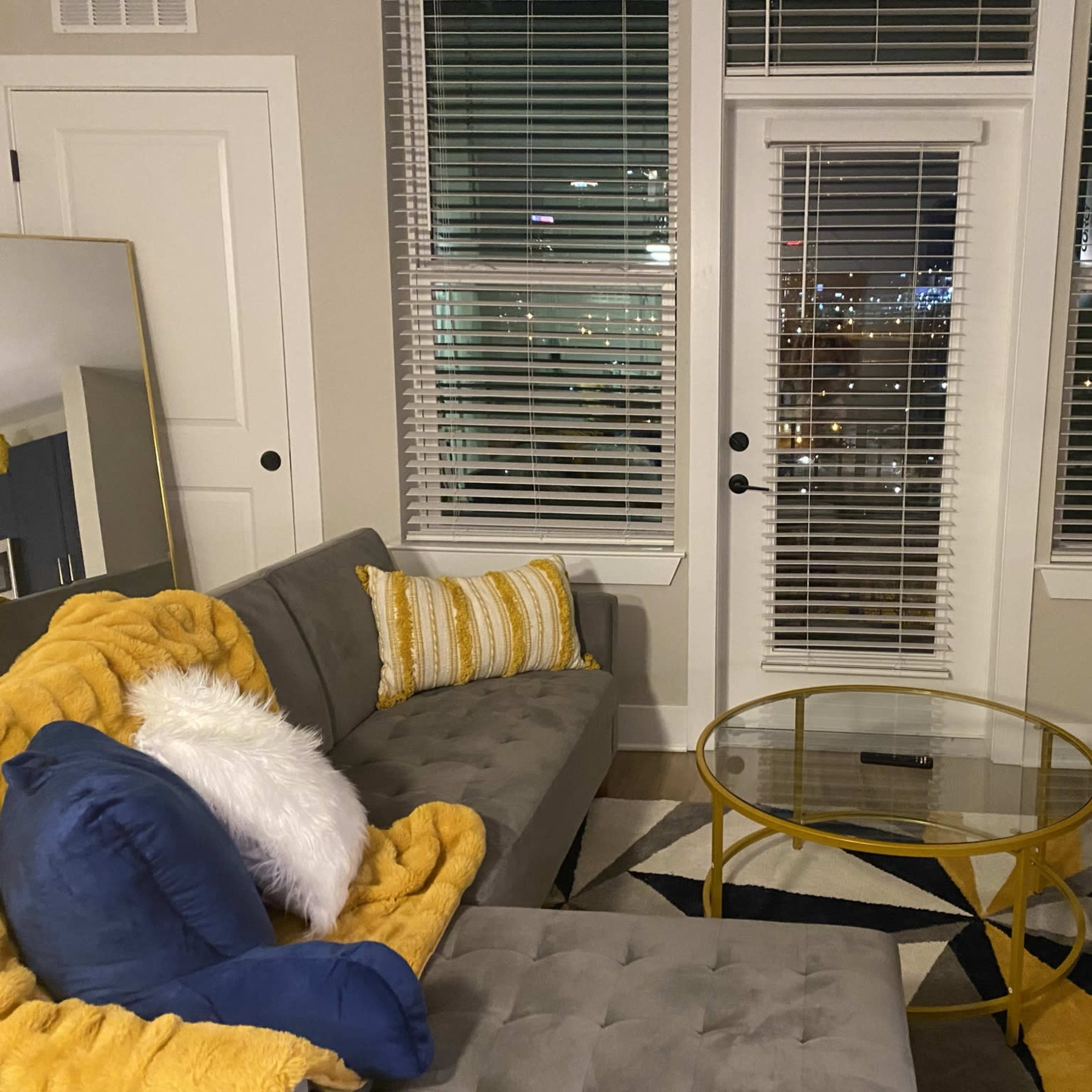 The room features a gray sectional sofa adorned with yellow and white pillows, a circular glass coffee table, and large windows that overlook a cityscape at night.