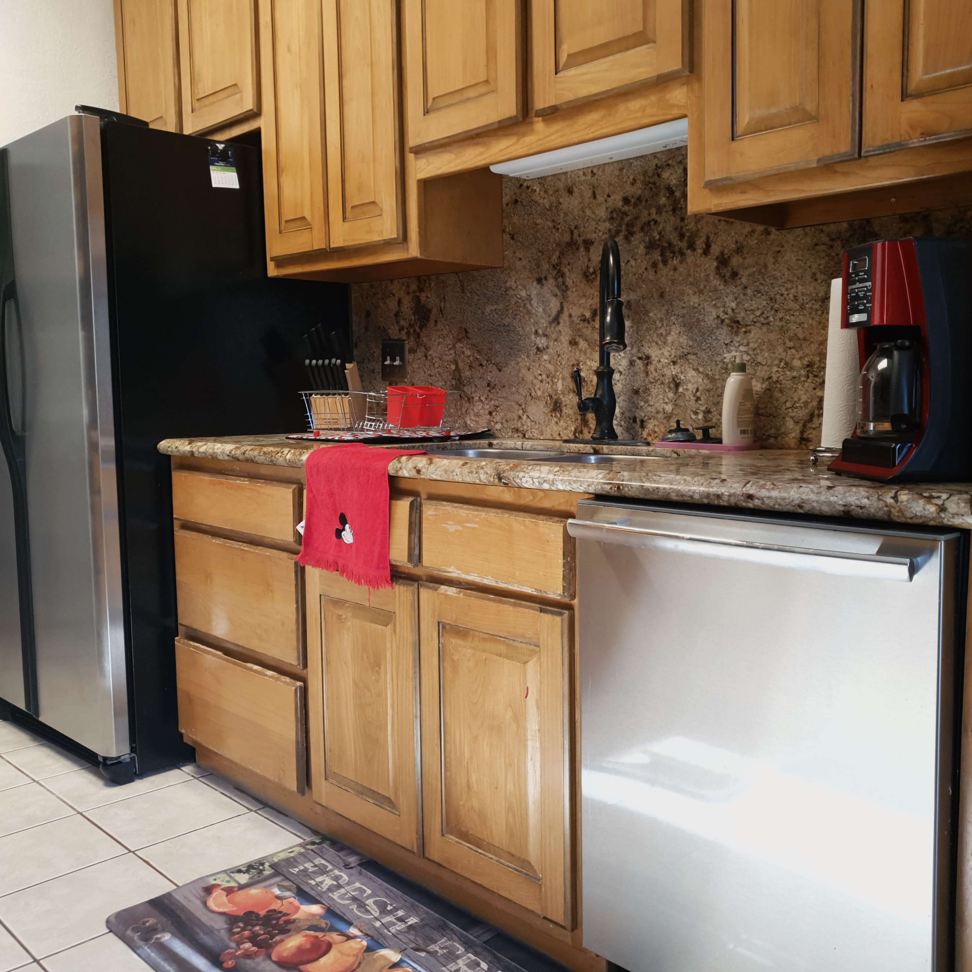 The kitchen features wooden cabinets, a stainless steel refrigerator, a coffee maker, and a dishwashing machine, all arranged along a tiled floor.