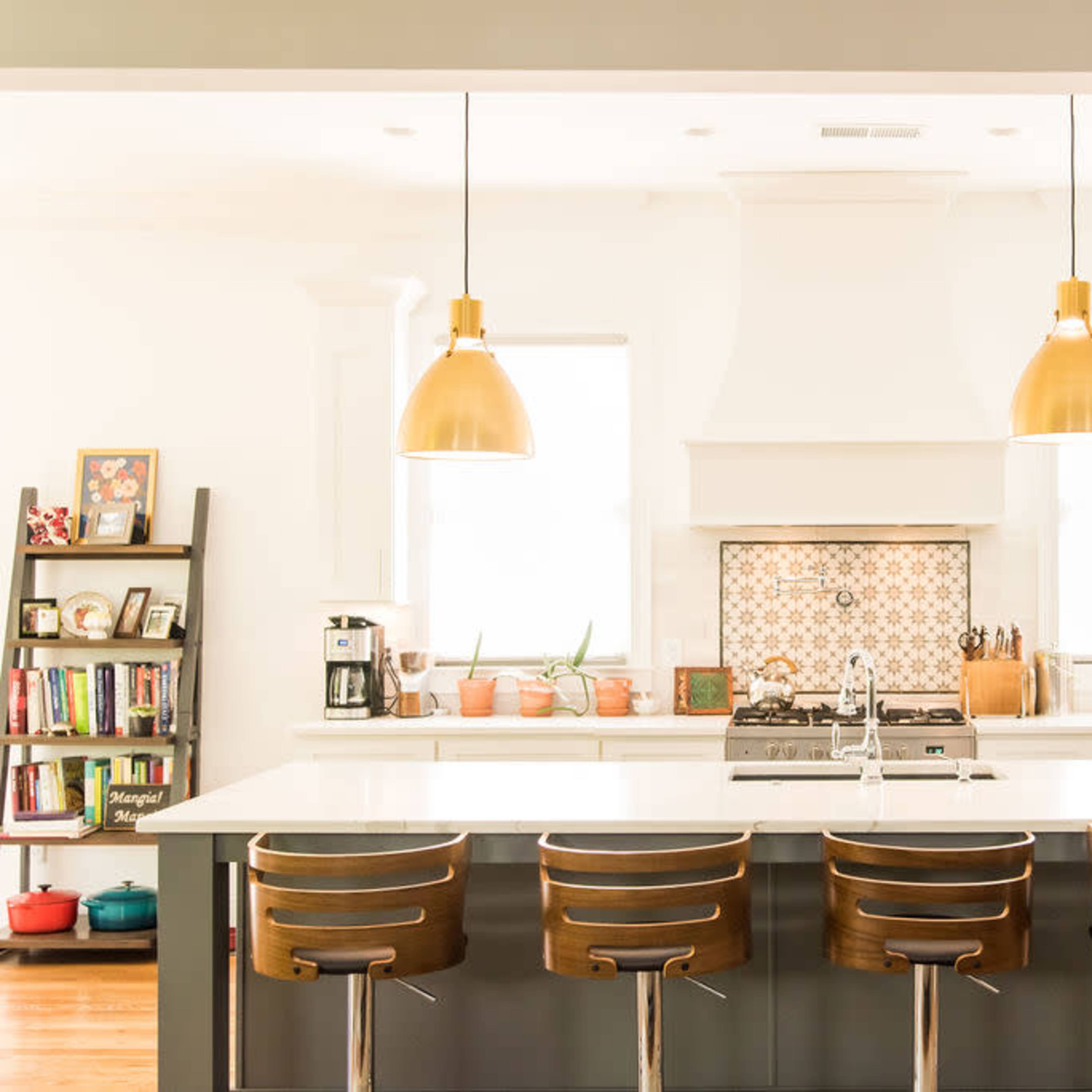 A modern kitchen features a large island with four wooden bar stools, pendant lights above, and a backsplash with a tile design, flanked by white cabinets and stainless steel appliances.