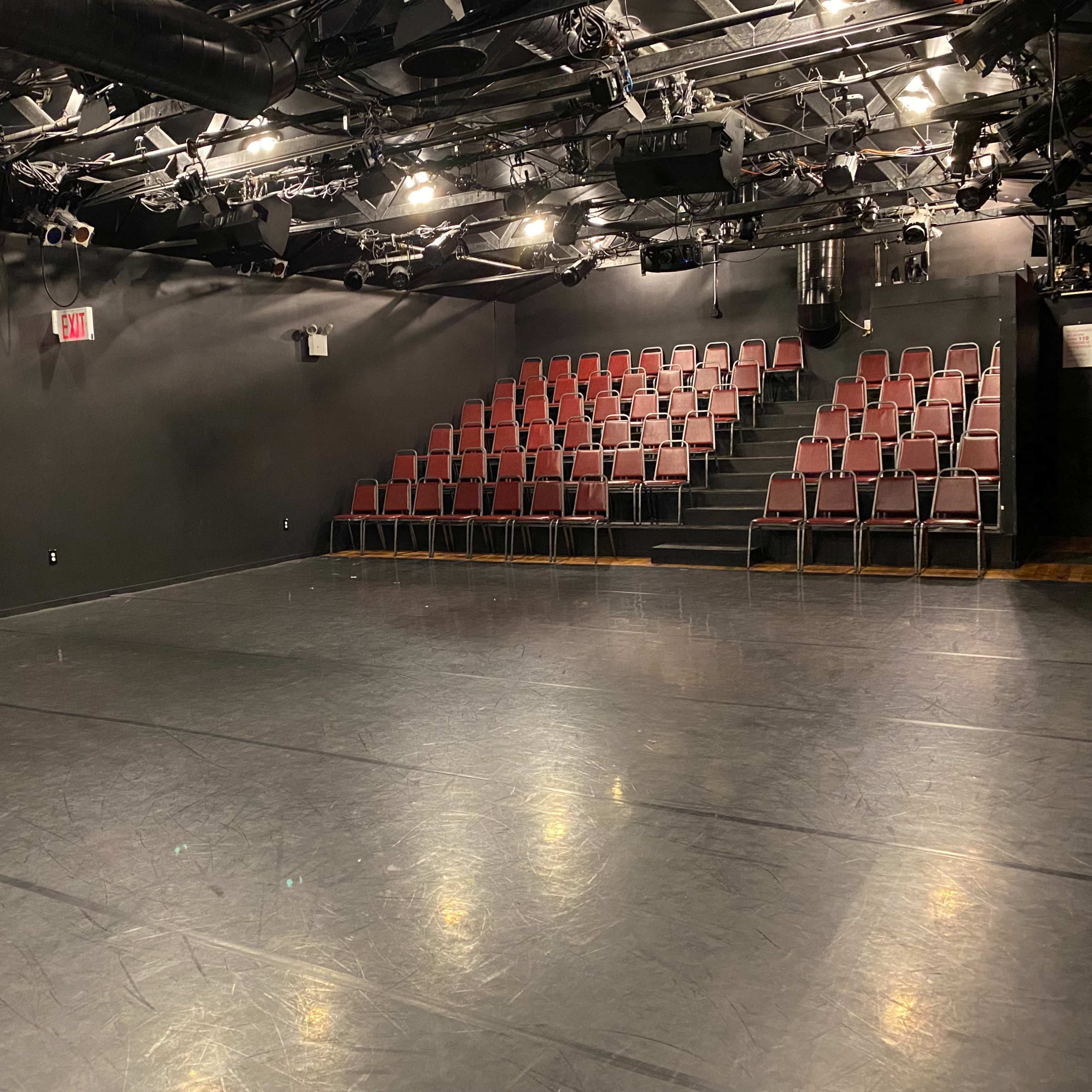 A darkened performance space with a flat, shiny floor and a row of red chairs arranged in a tiered seating area.