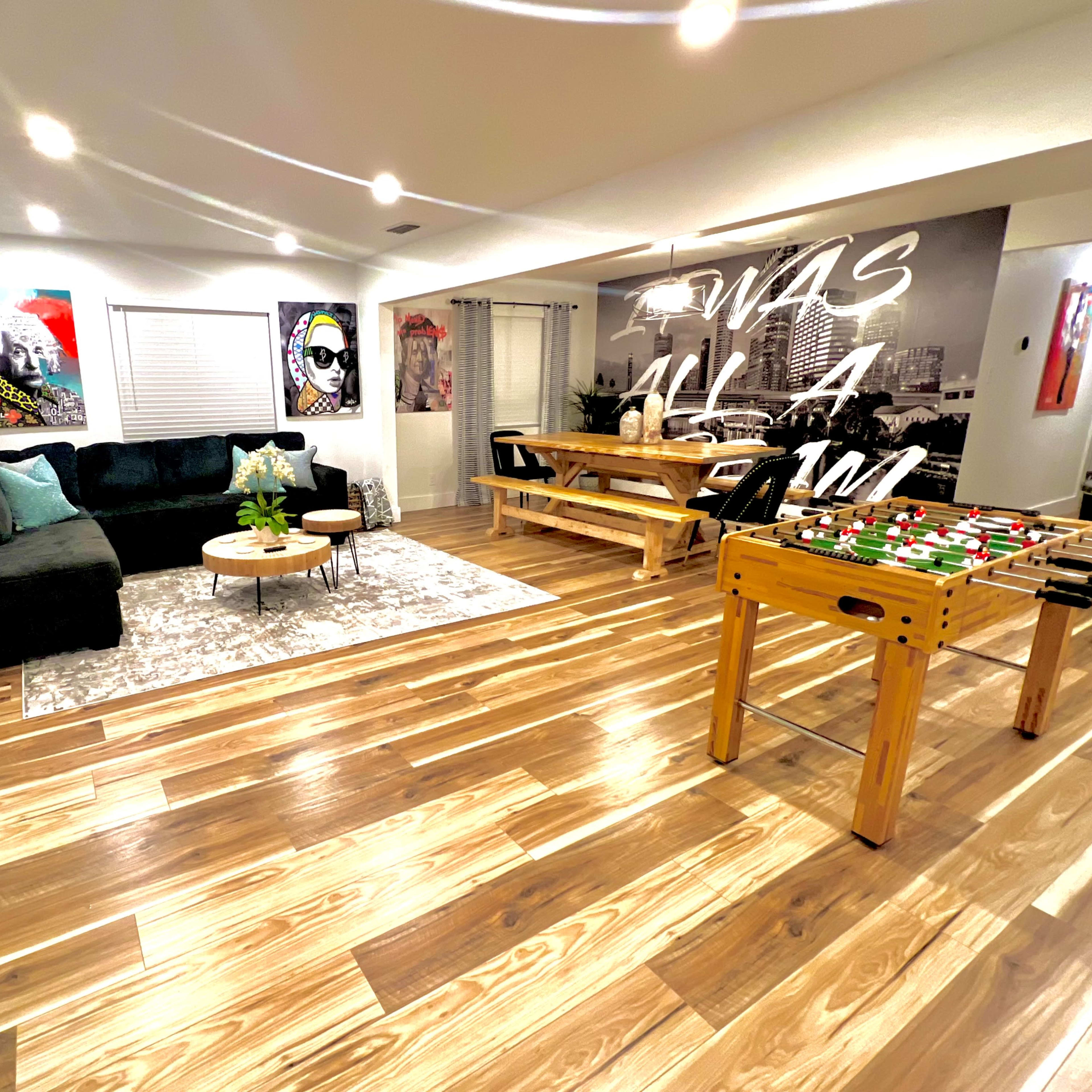 The room features a dark sofa, a round coffee table, a foosball table, and dining area with artwork on the walls and wooden flooring.
