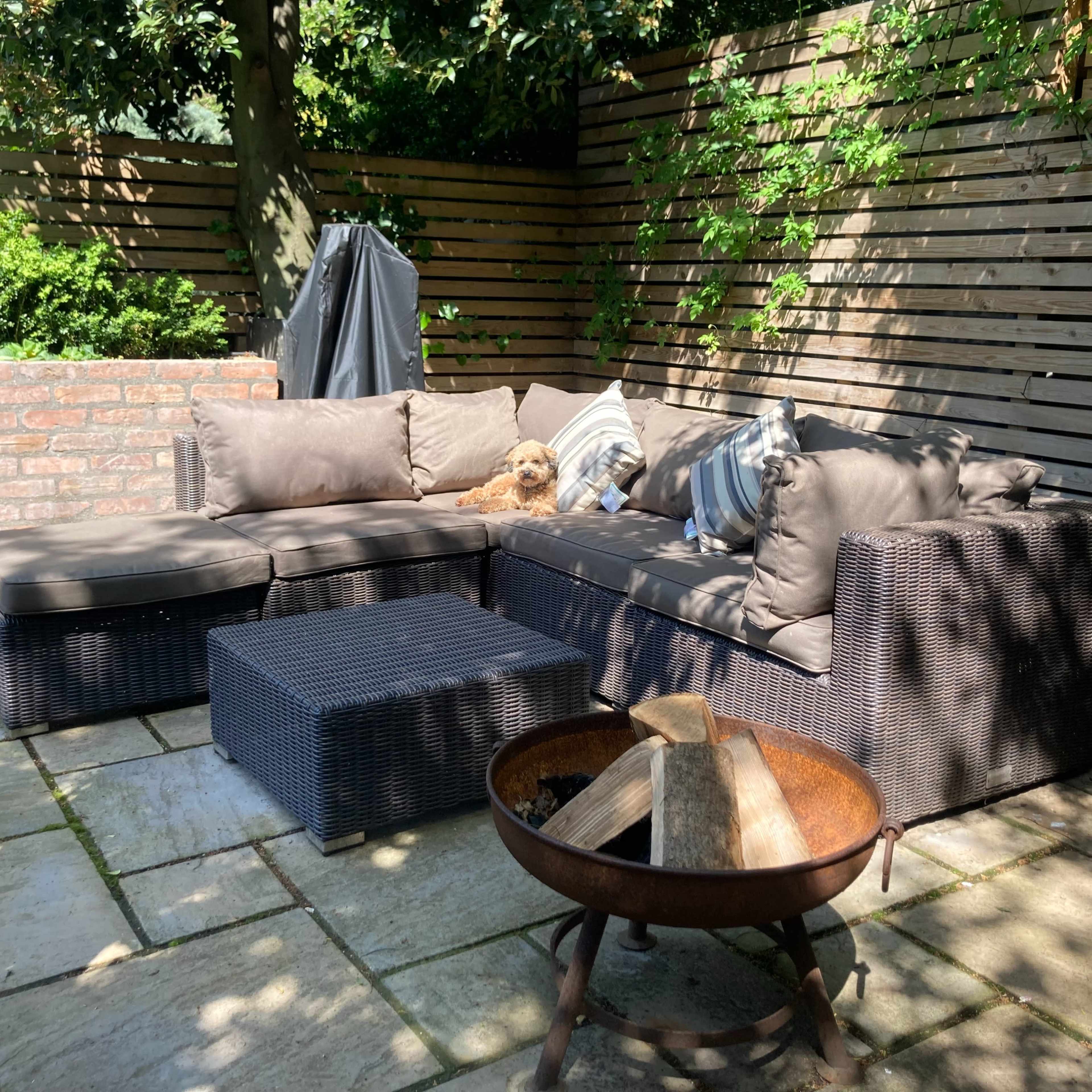 A patio area features a brown wicker sectional sofa with cushions, a matching coffee table, and a metal fire pit surrounded by stone tiles, with a small dog resting on the sofa.
