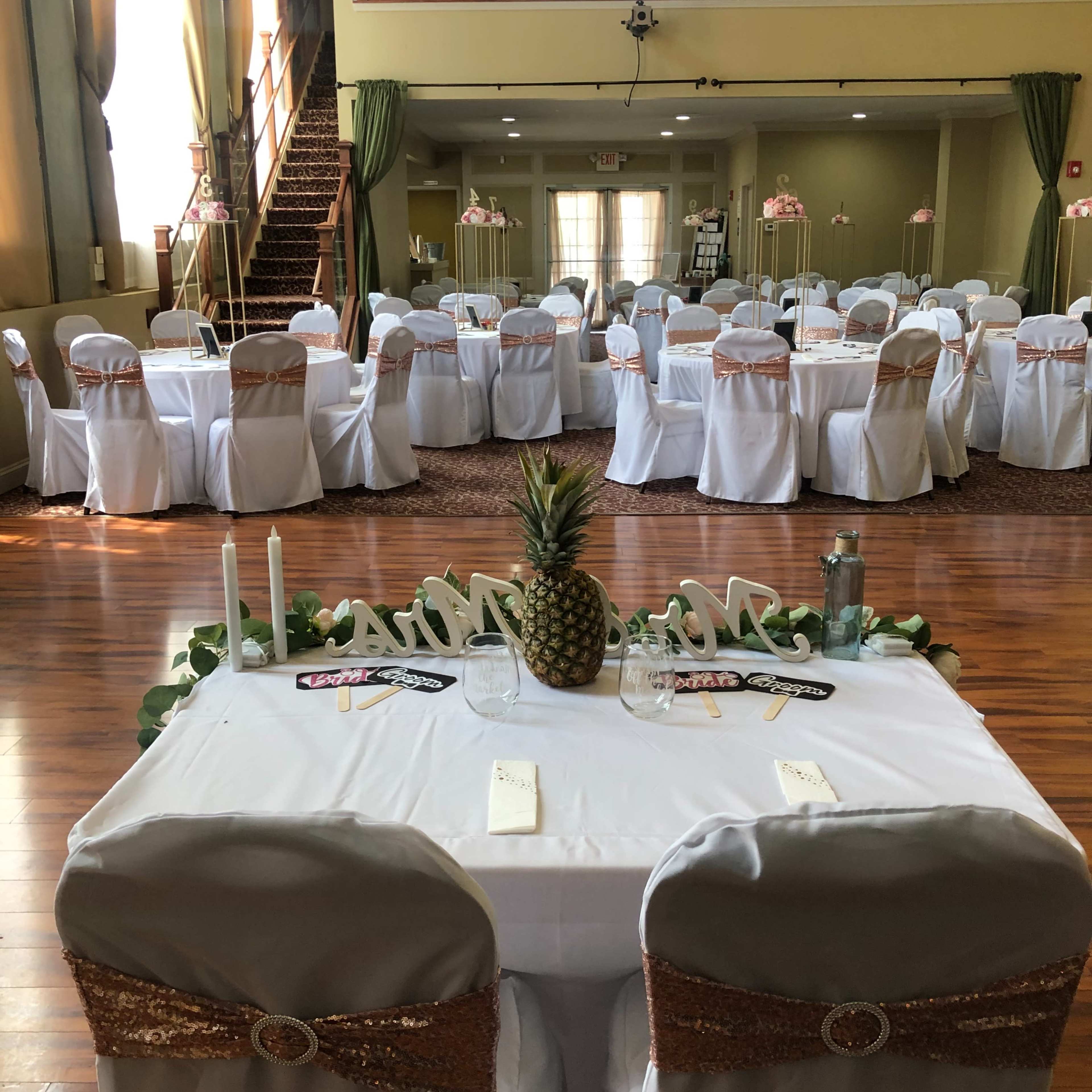 A banquet hall is set up for an event, featuring several tables covered with white tablecloths and decorated with rose-gold accents.