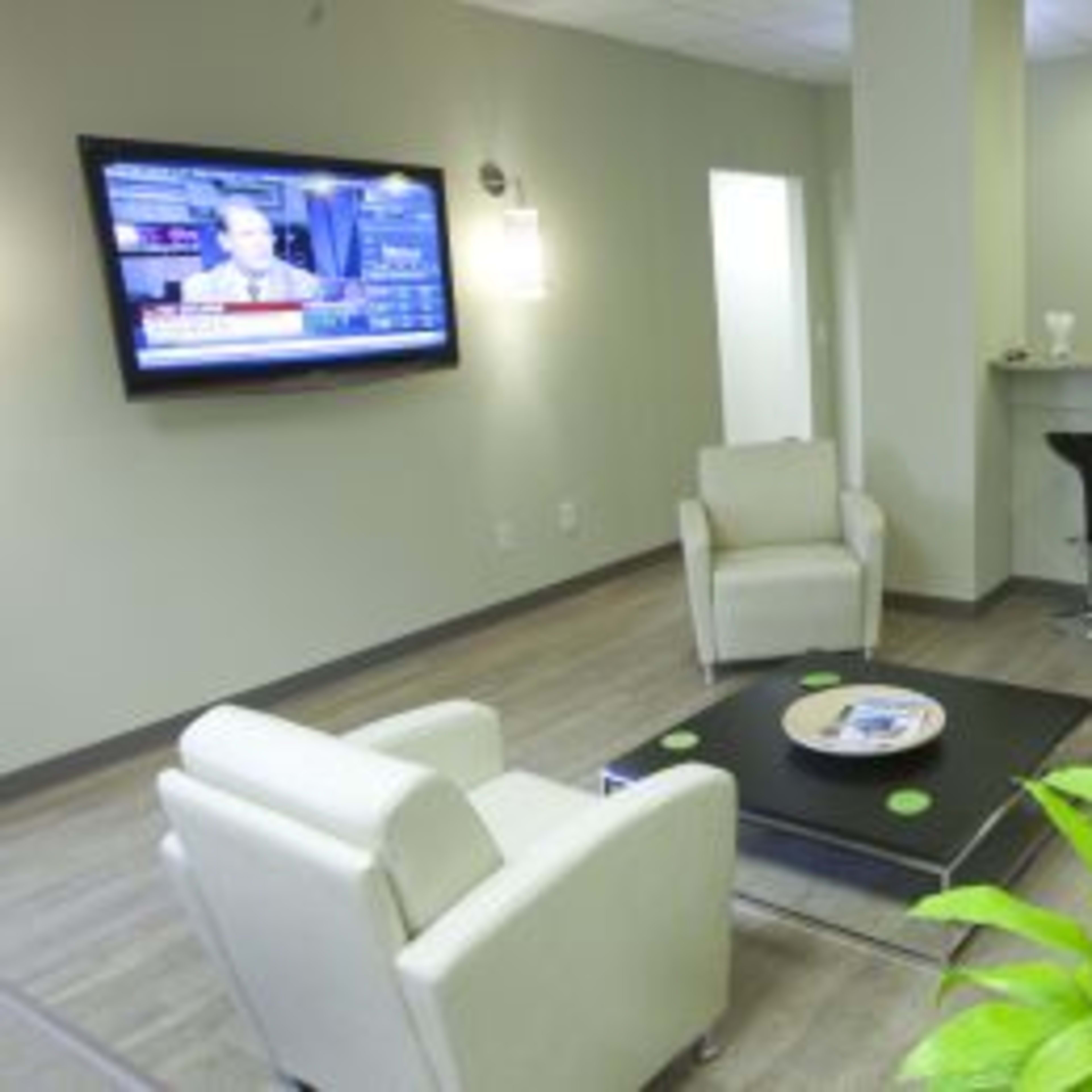 A modern waiting area with two white armchairs, a coffee table, a plant, a flat-screen TV displaying news, and a small bar with high stools.